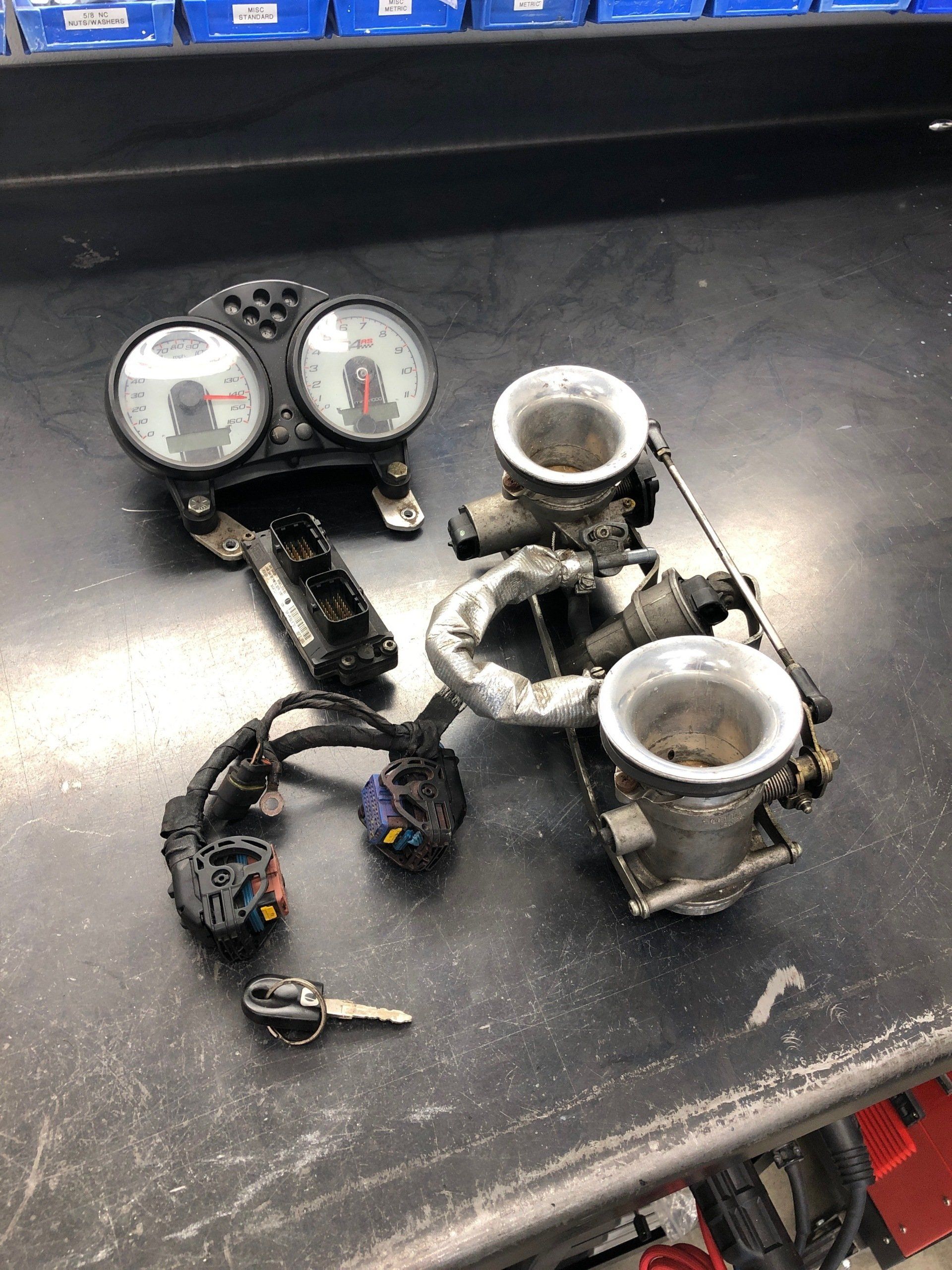 Disassembled motorcycle parts on a black workbench, including gauges, clamps, wiring, and a key.