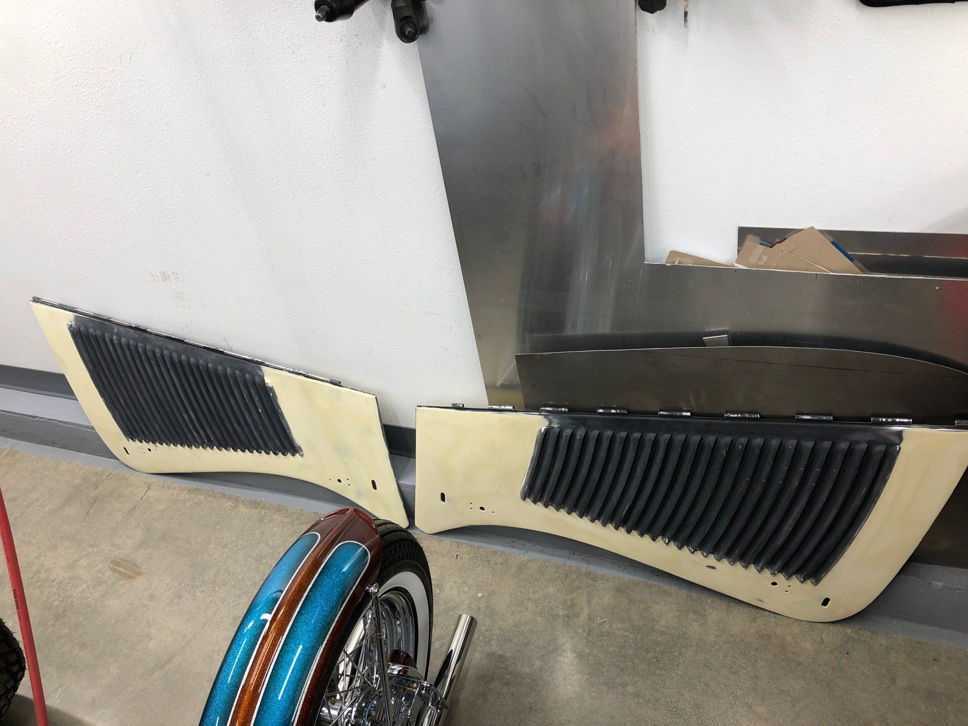 Broken beige car body panel with black vents leaning on a garage floor рядом with tools and blue hose reel