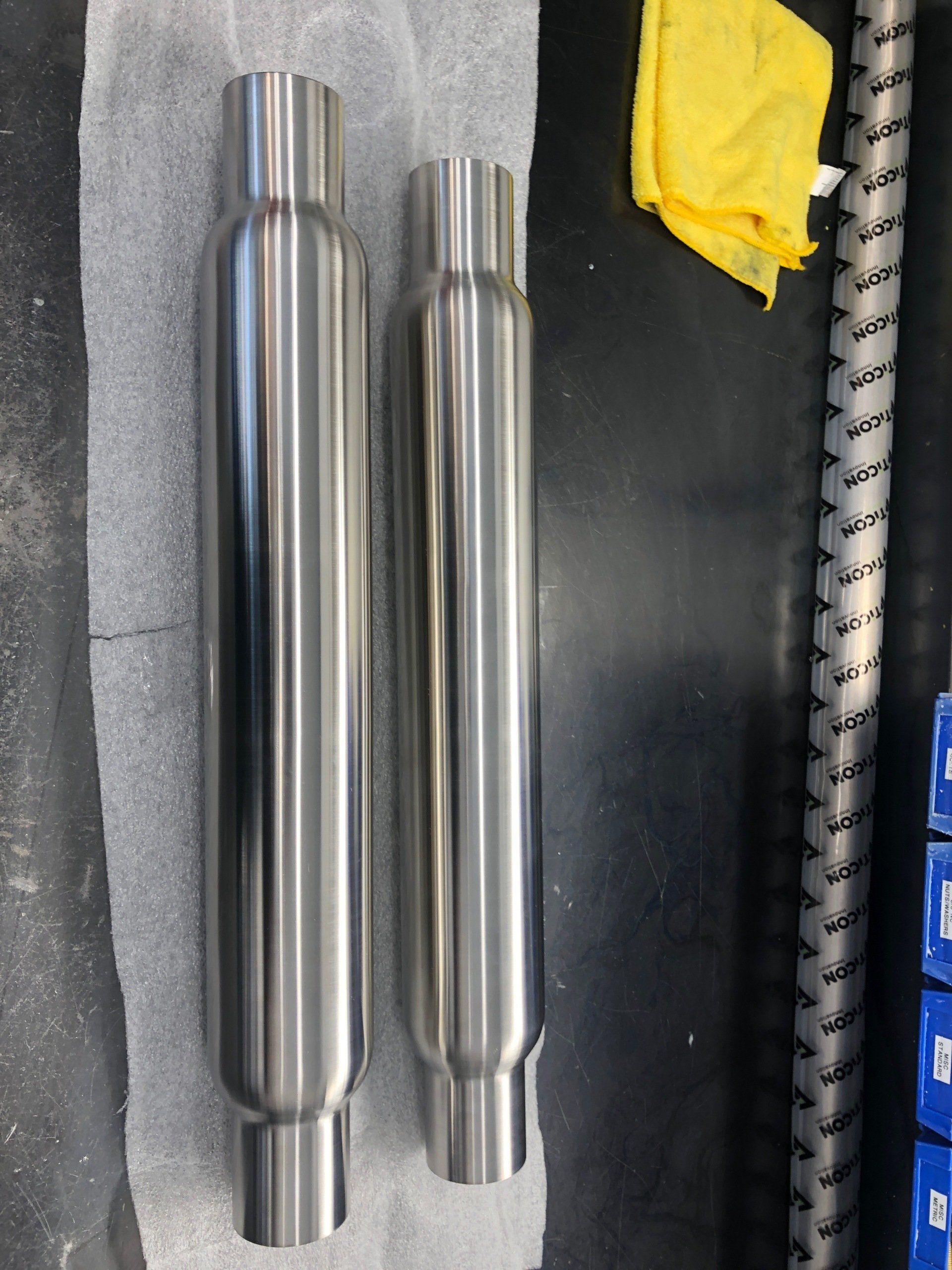 Two shiny metal cylindrical parts on a dark workbench beside a yellow cloth and a coiled cable.