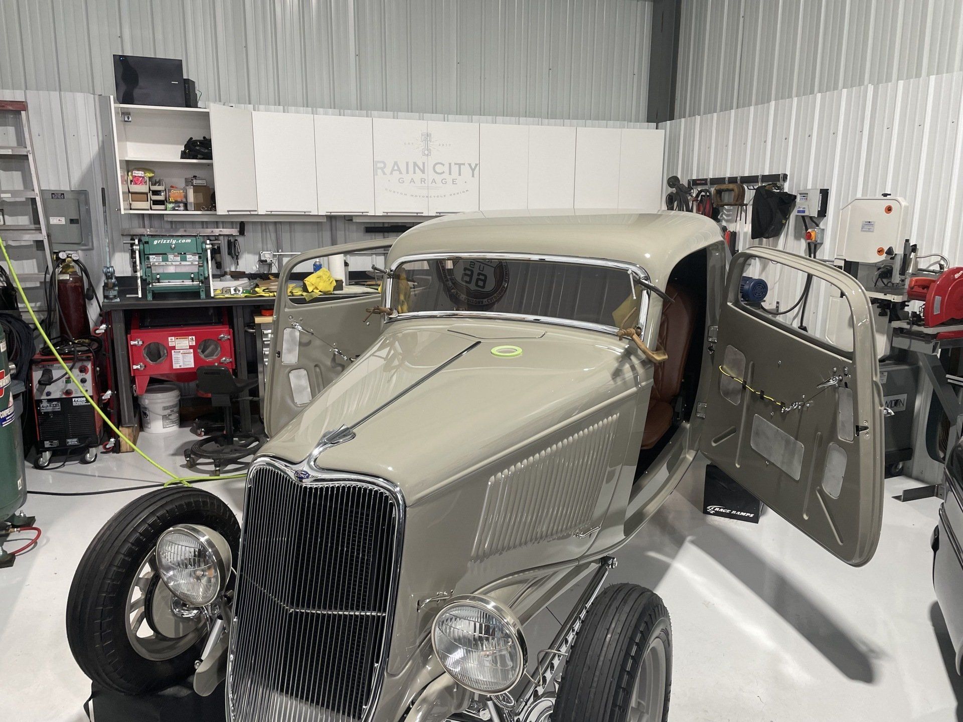 Vintage gray car in a garage with the hood open and doors ajar, surrounded by tools and equipment.