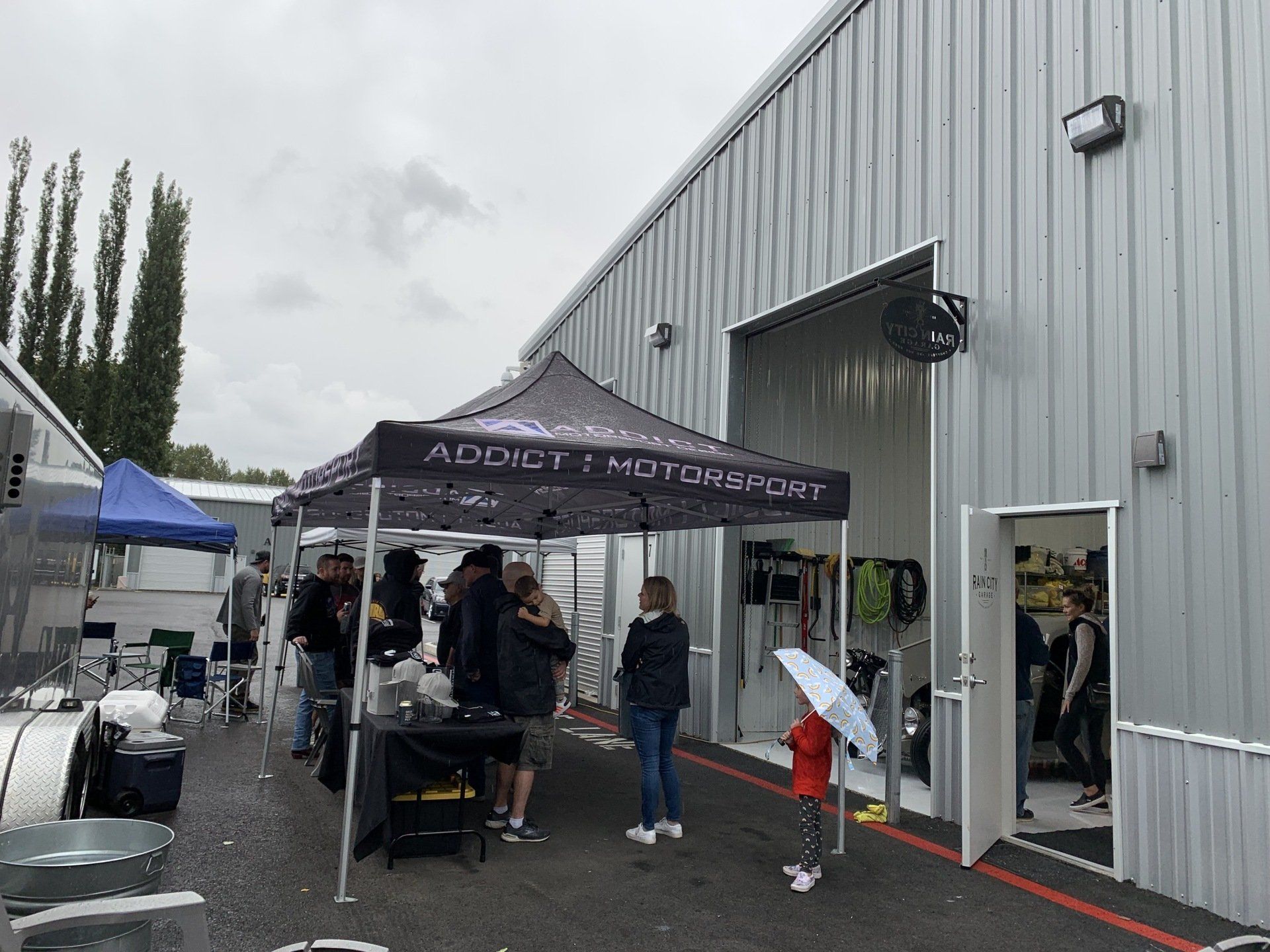 Outdoor market tent by a gray warehouse, with people gathered under a black canopy on a wet pavement.