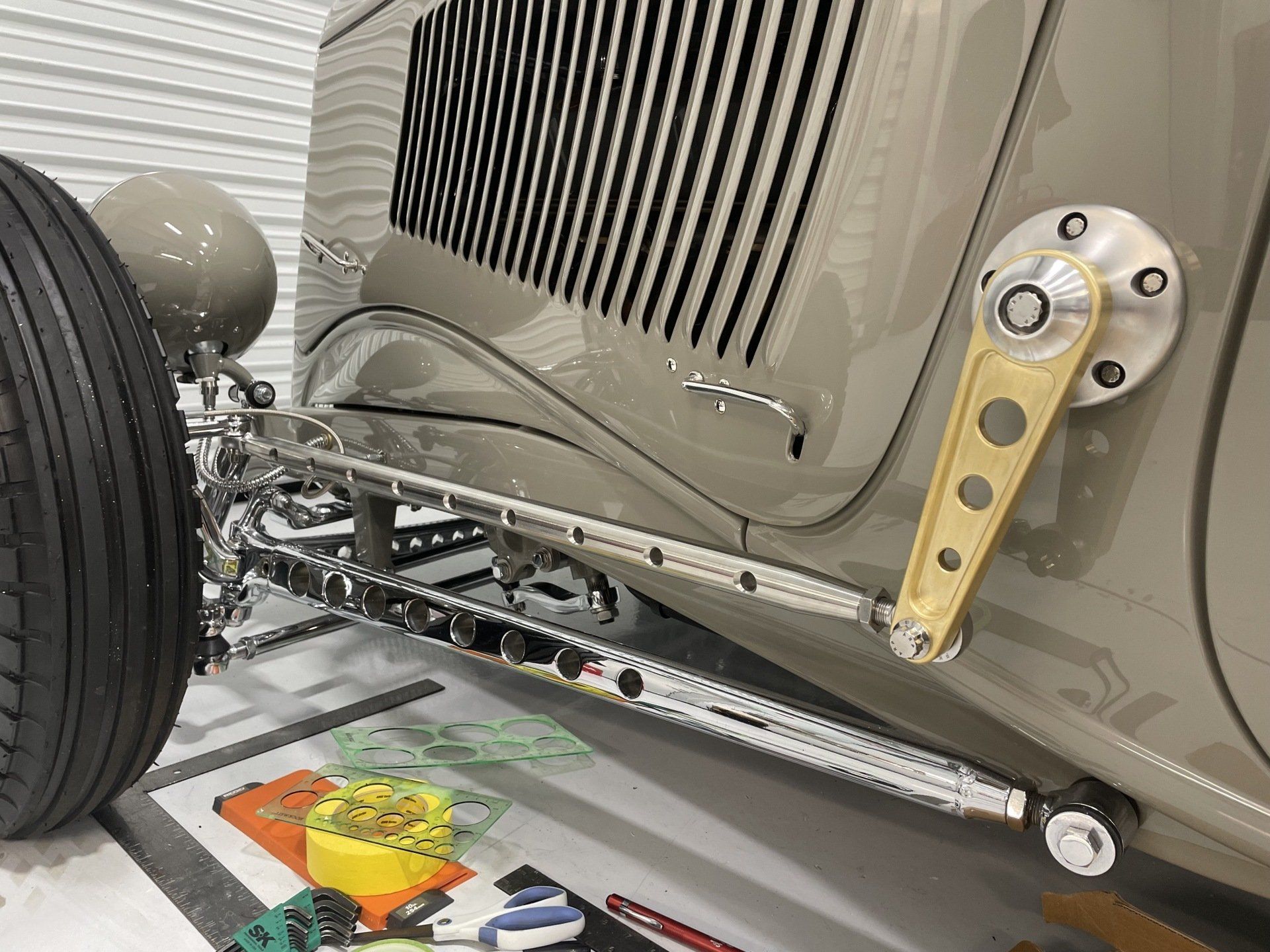 Close-up of a vintage car chassis and suspension assembly in a workshop, with front wheel and chrome steering linkage visible