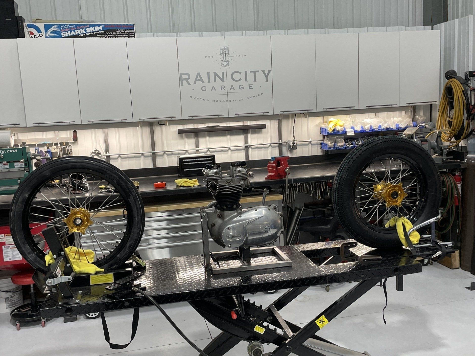 Race car chassis on a lift in a workshop, with four wheels attached and a Rain City banner in the background