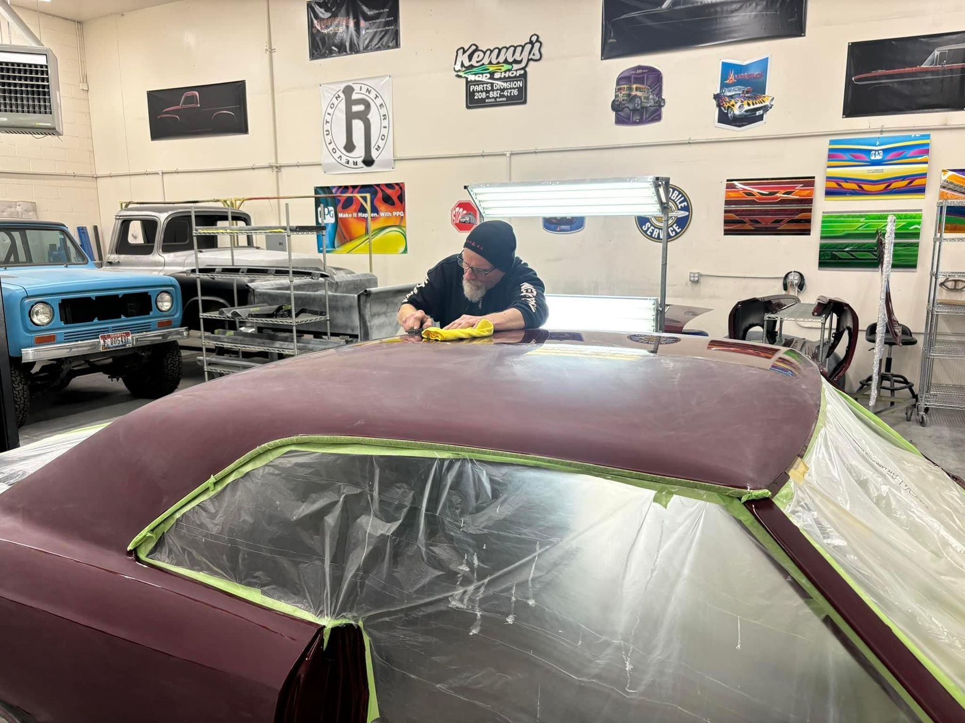 Person working on a maroon car in a garage, surrounded by tools and classic vehicles.