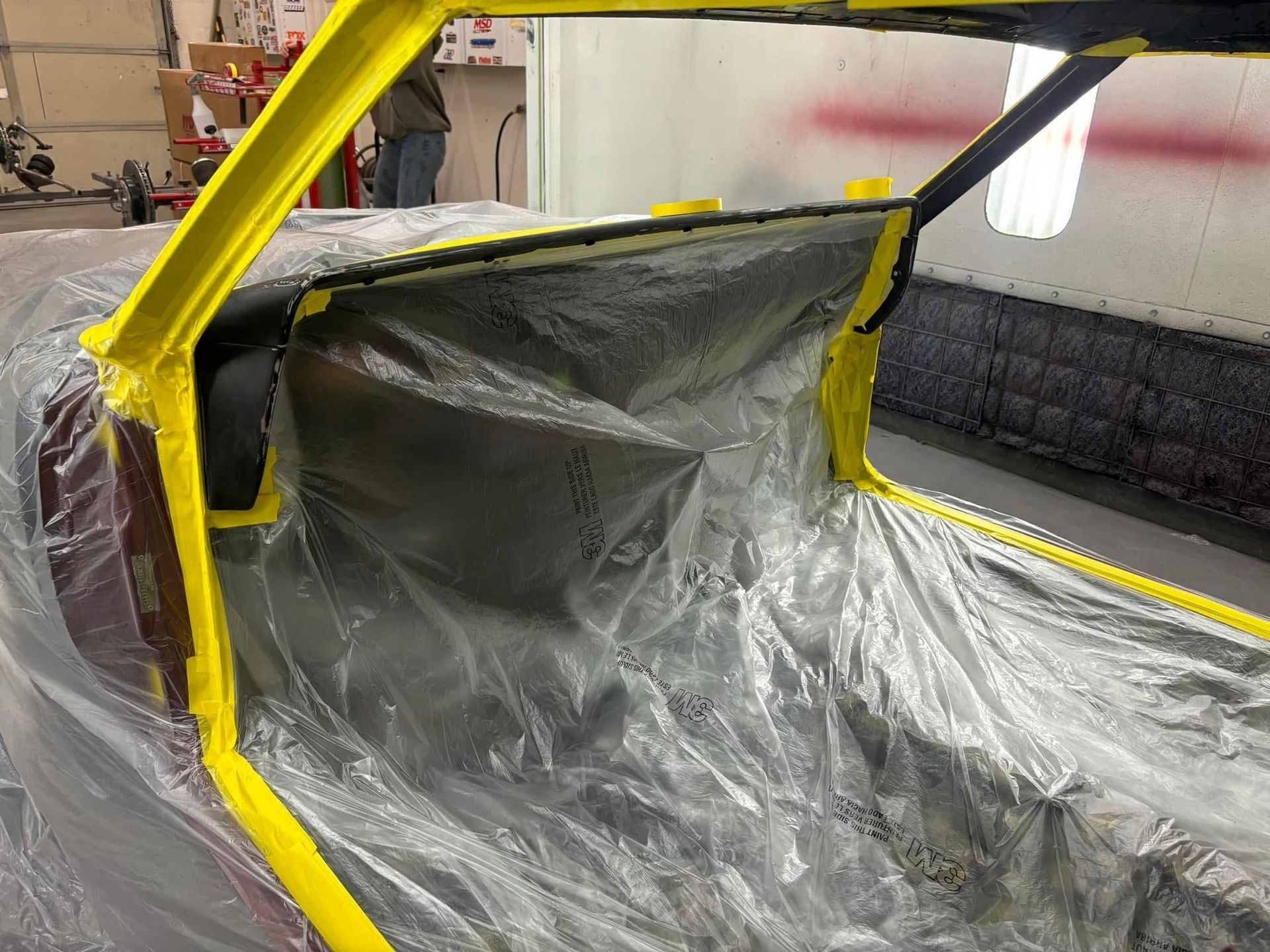 Yellow car body covered in plastic sheeting inside a workshop, likely for painting or repair