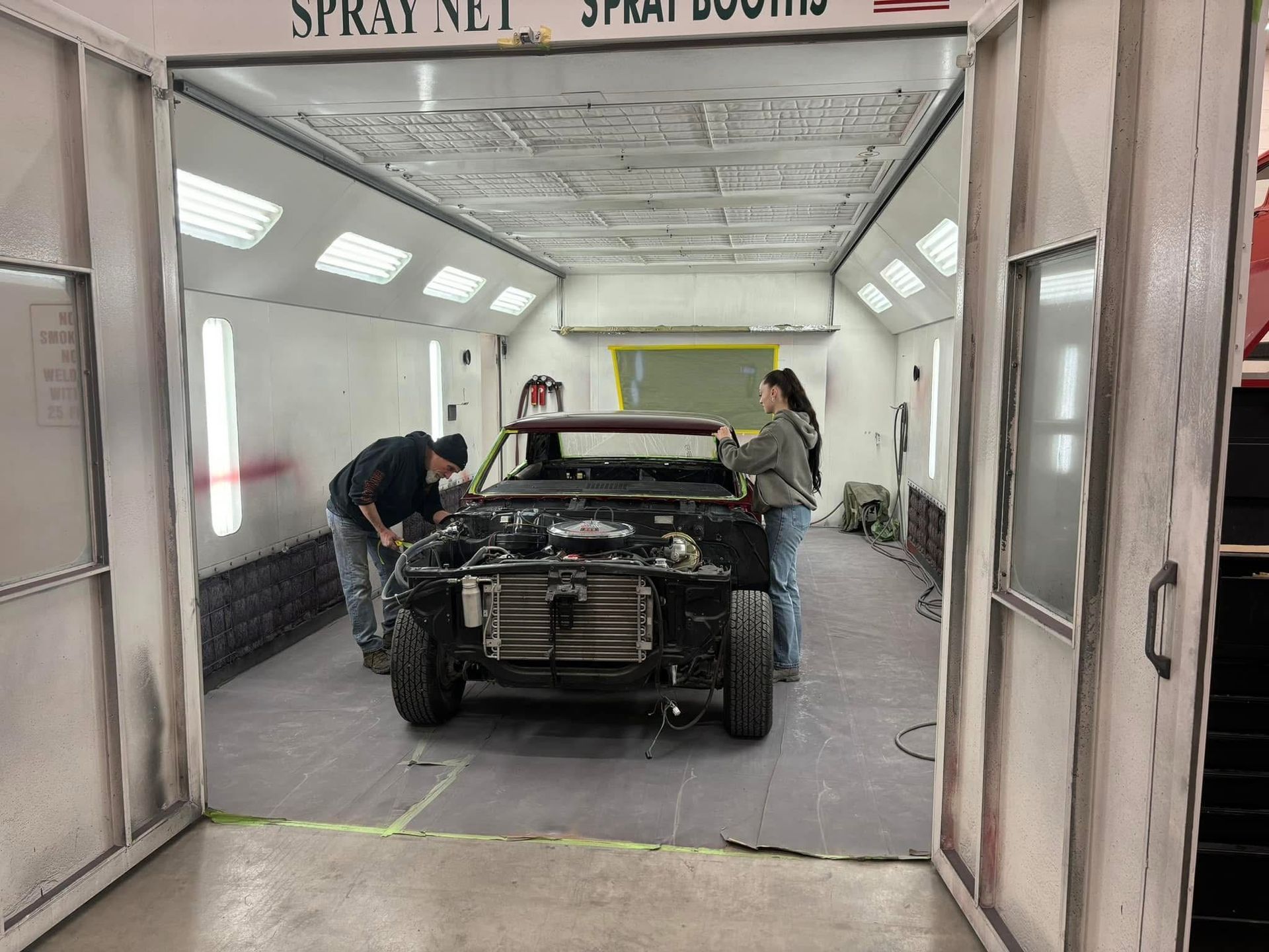 Car being repaired in a spray booth, with two mechanics working on it.