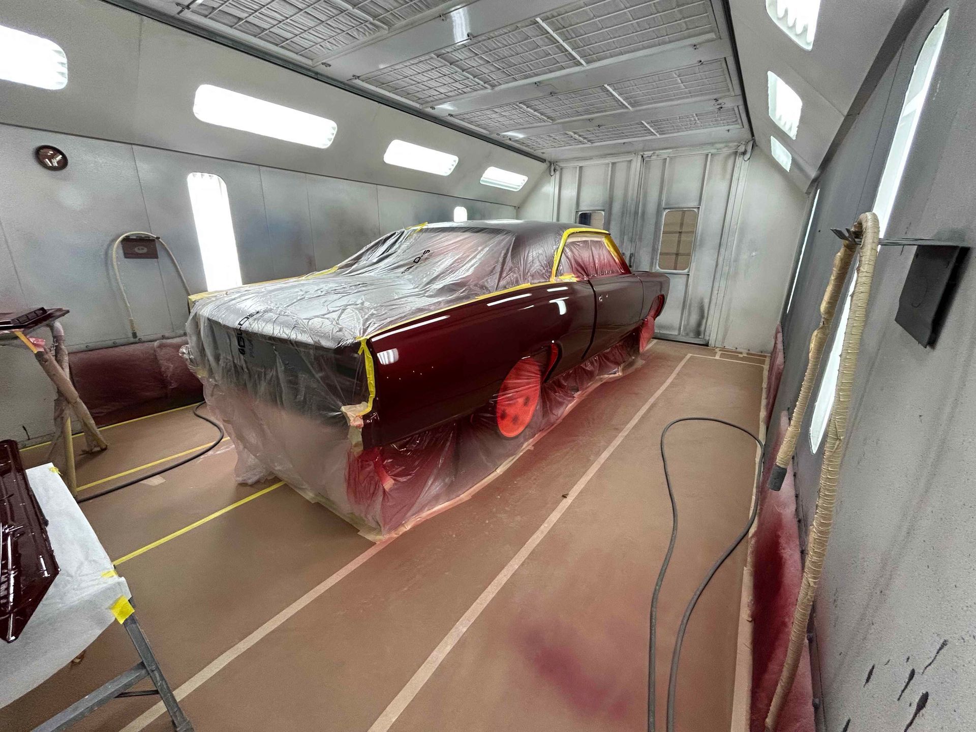 Classic car masked and primed in a spray booth, with red wheels and plastic sheeting covering the body.