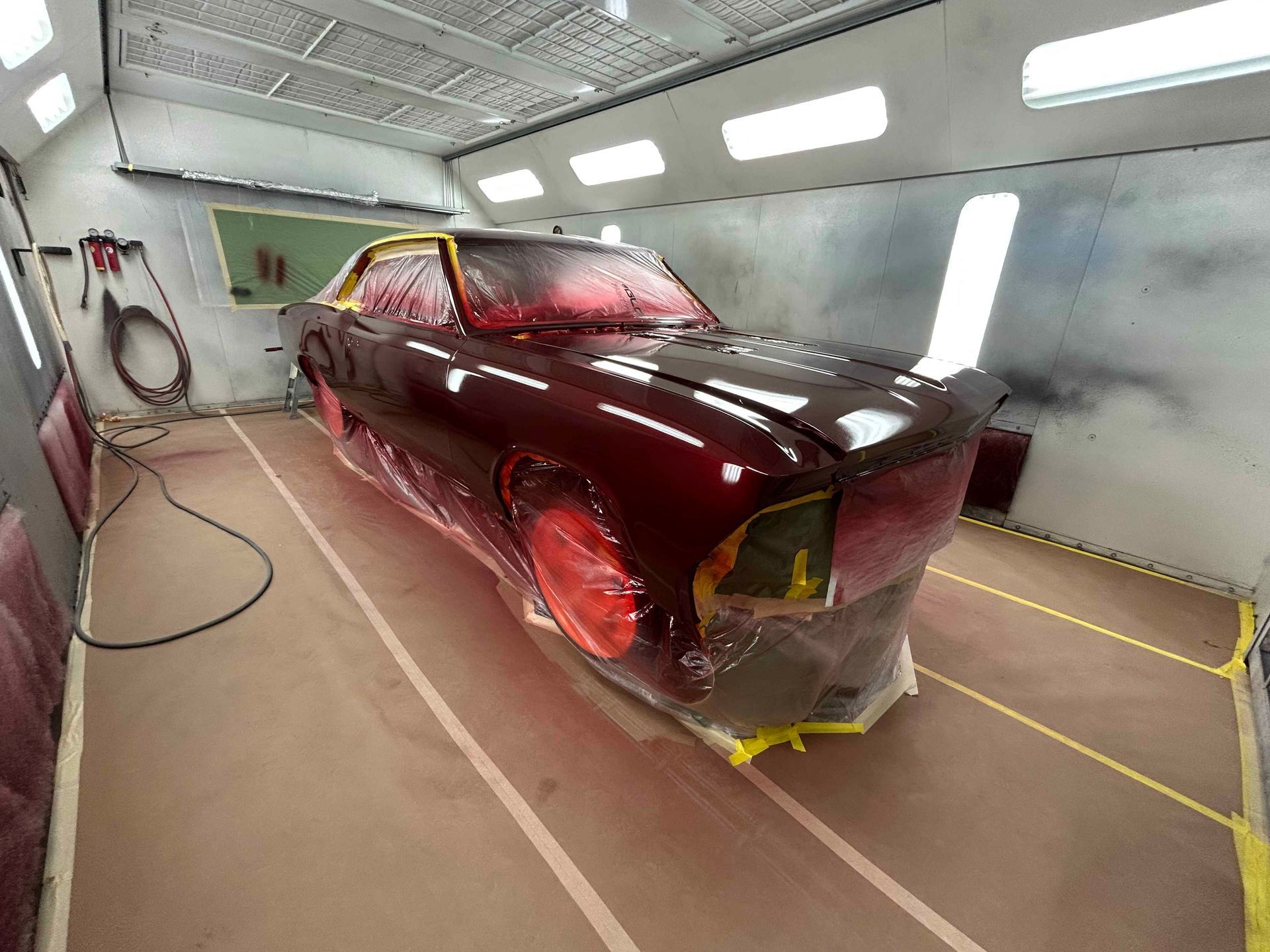 Maroon car body in a paint booth, covered in primer or paint, with masking and spray equipment nearby