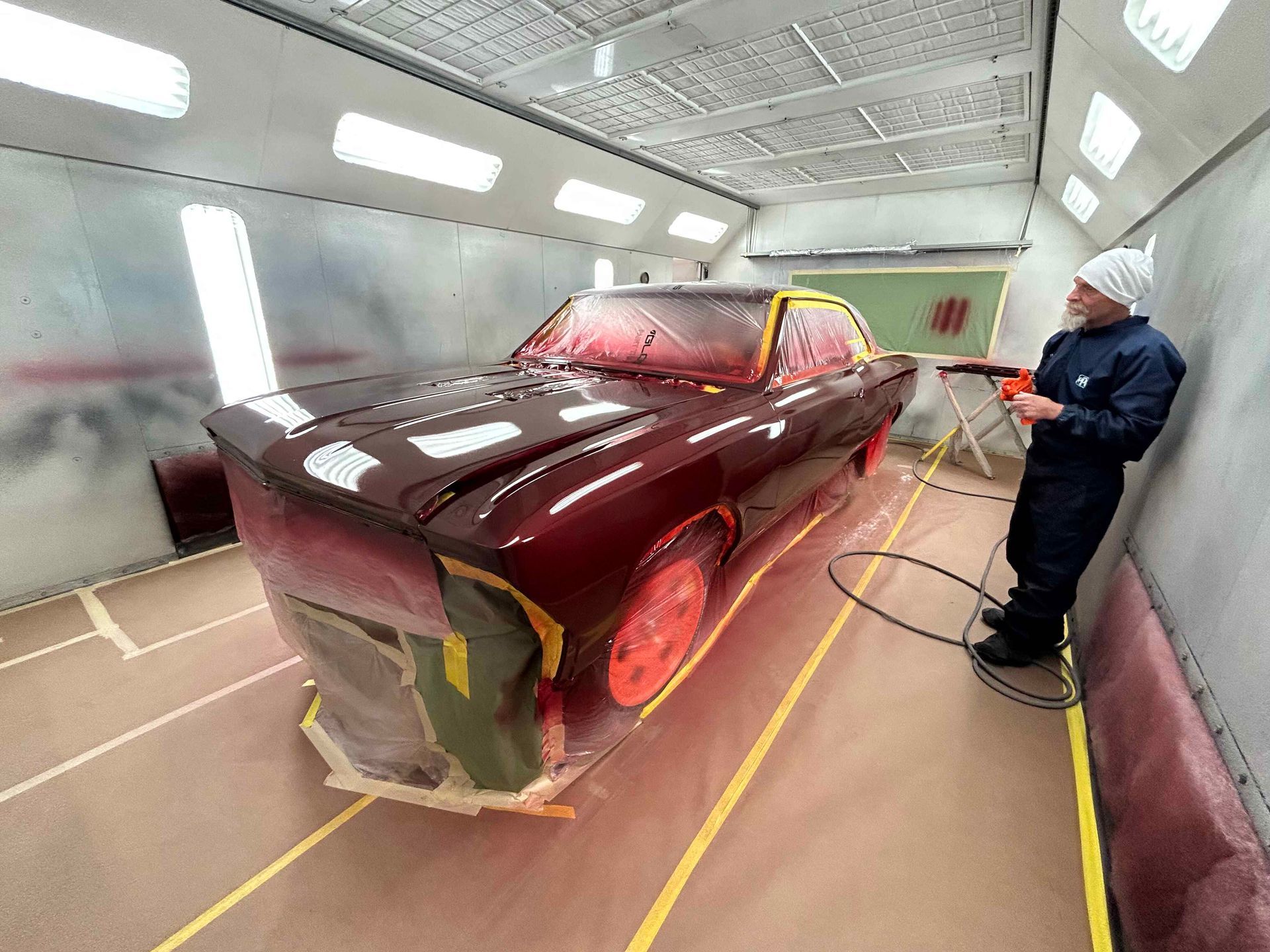 Maroon car in a paint booth while a worker sprays paint with a hose