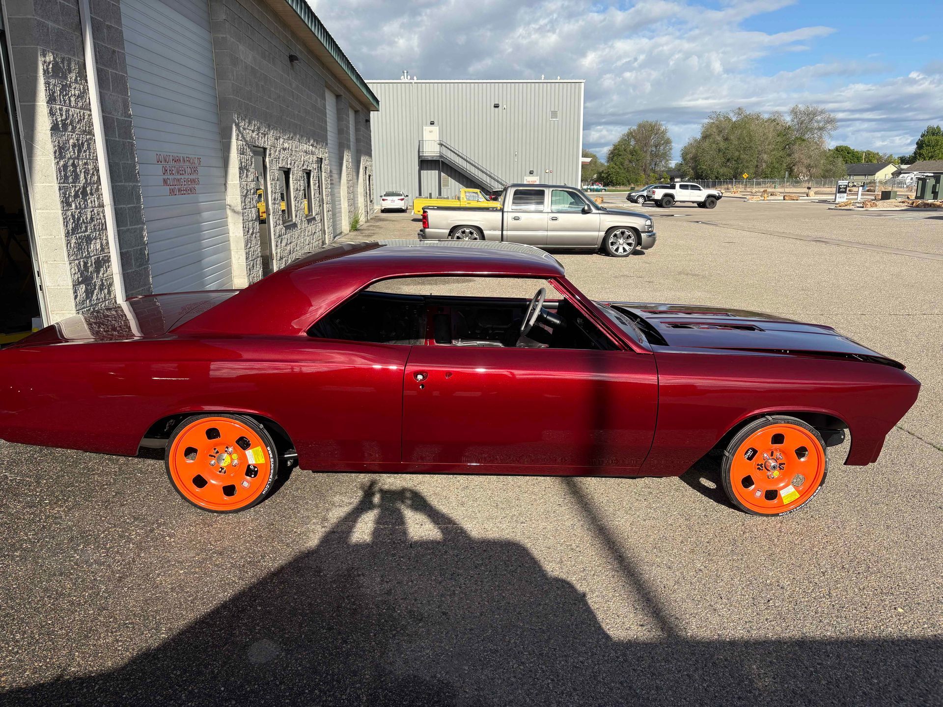 Maroon classic coupe with bright orange wheels parked beside a building on a gravel lot