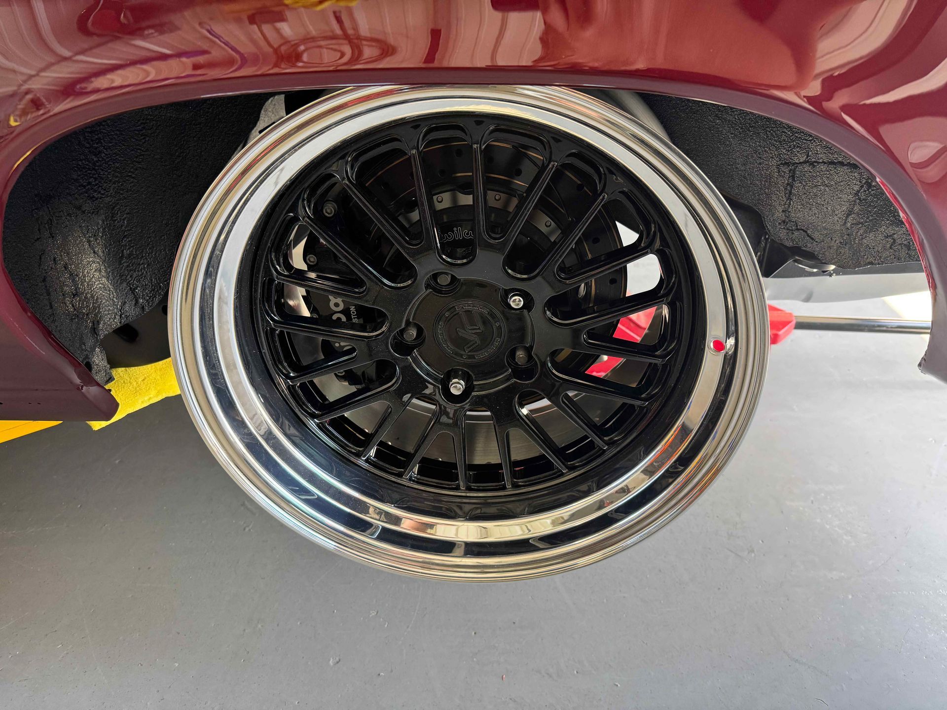 Close-up of a black custom wheel with a chrome lip on a maroon car, parked over a yellow curb