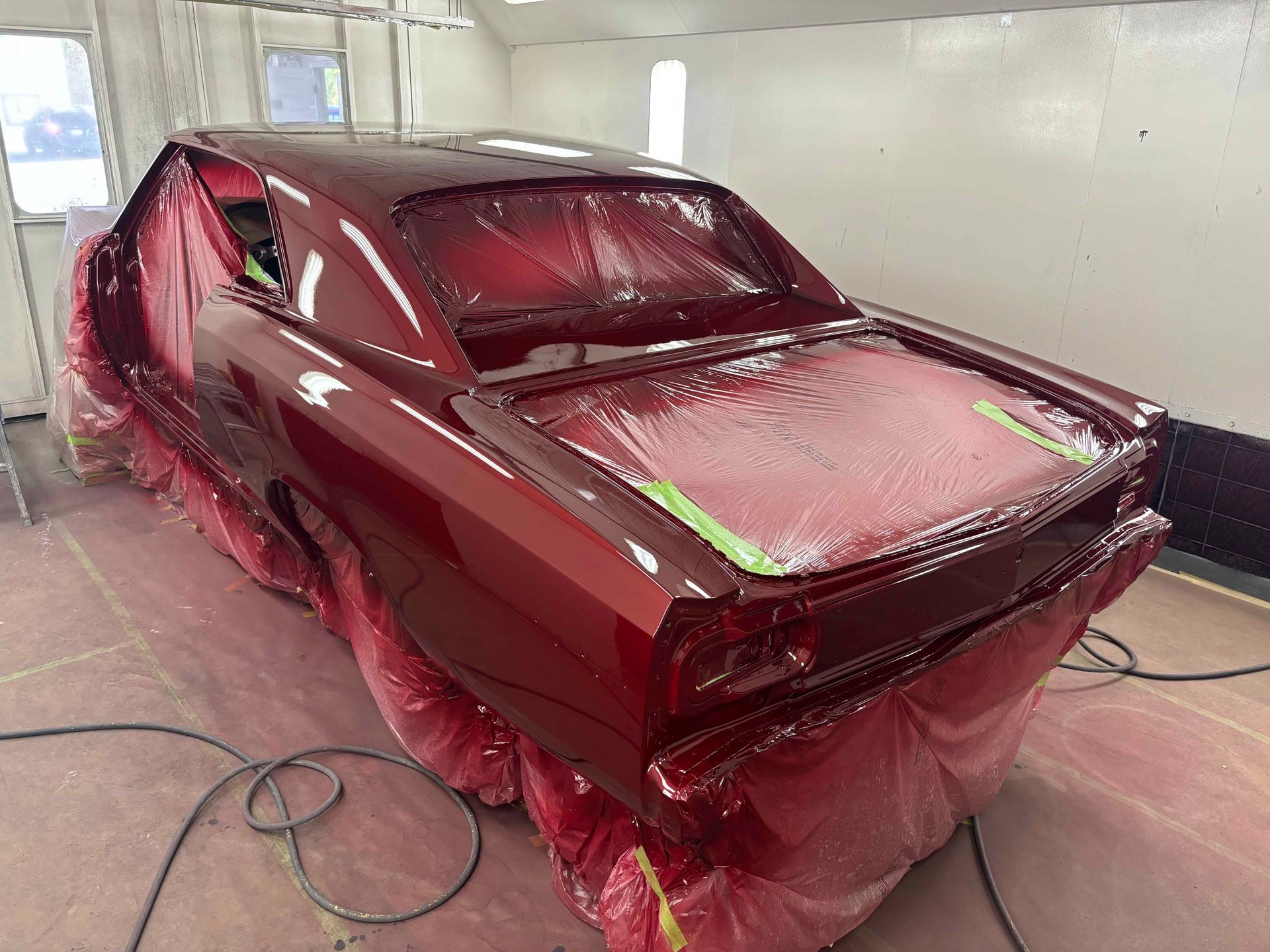 Red car body in a paint booth, partially masked with plastic sheeting.