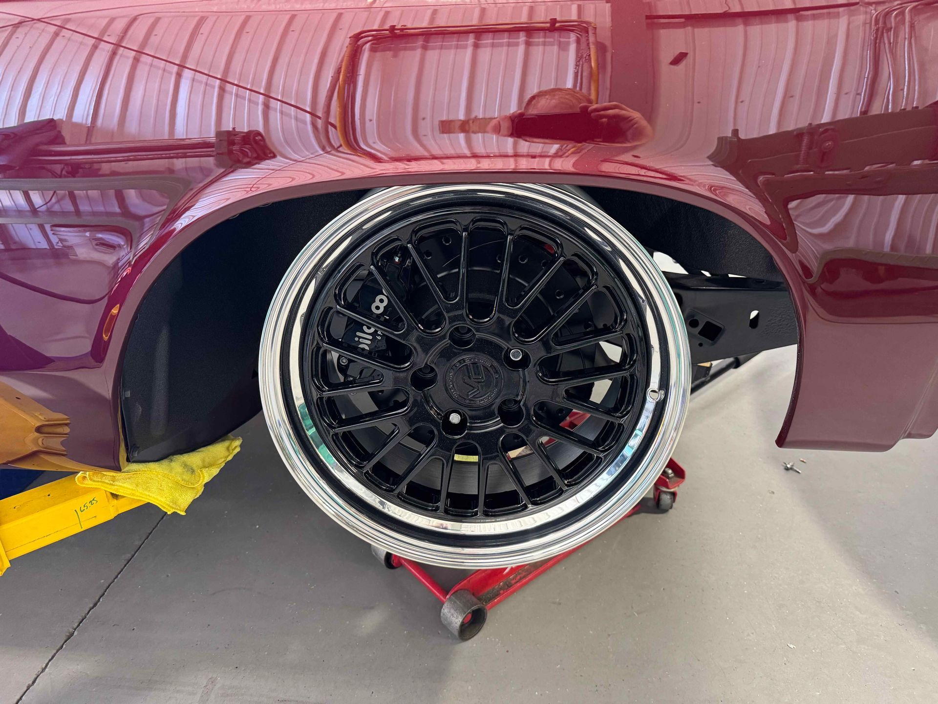 Maroon car rear wheel removed, showing black rim and brake assembly in a garage on a jack.