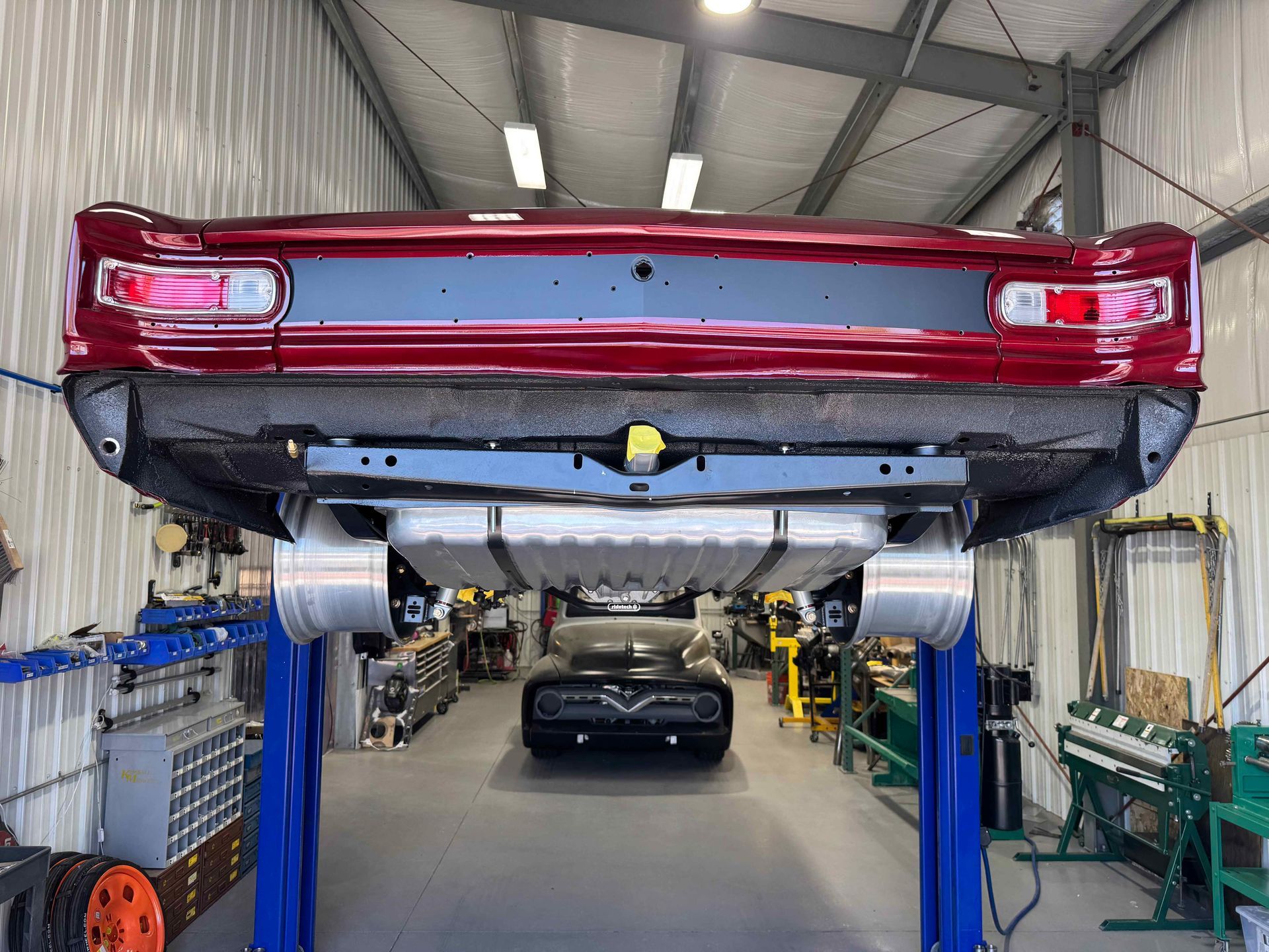 Red classic car lifted on a blue automotive hoist in a garage workshop with tools and another car in back