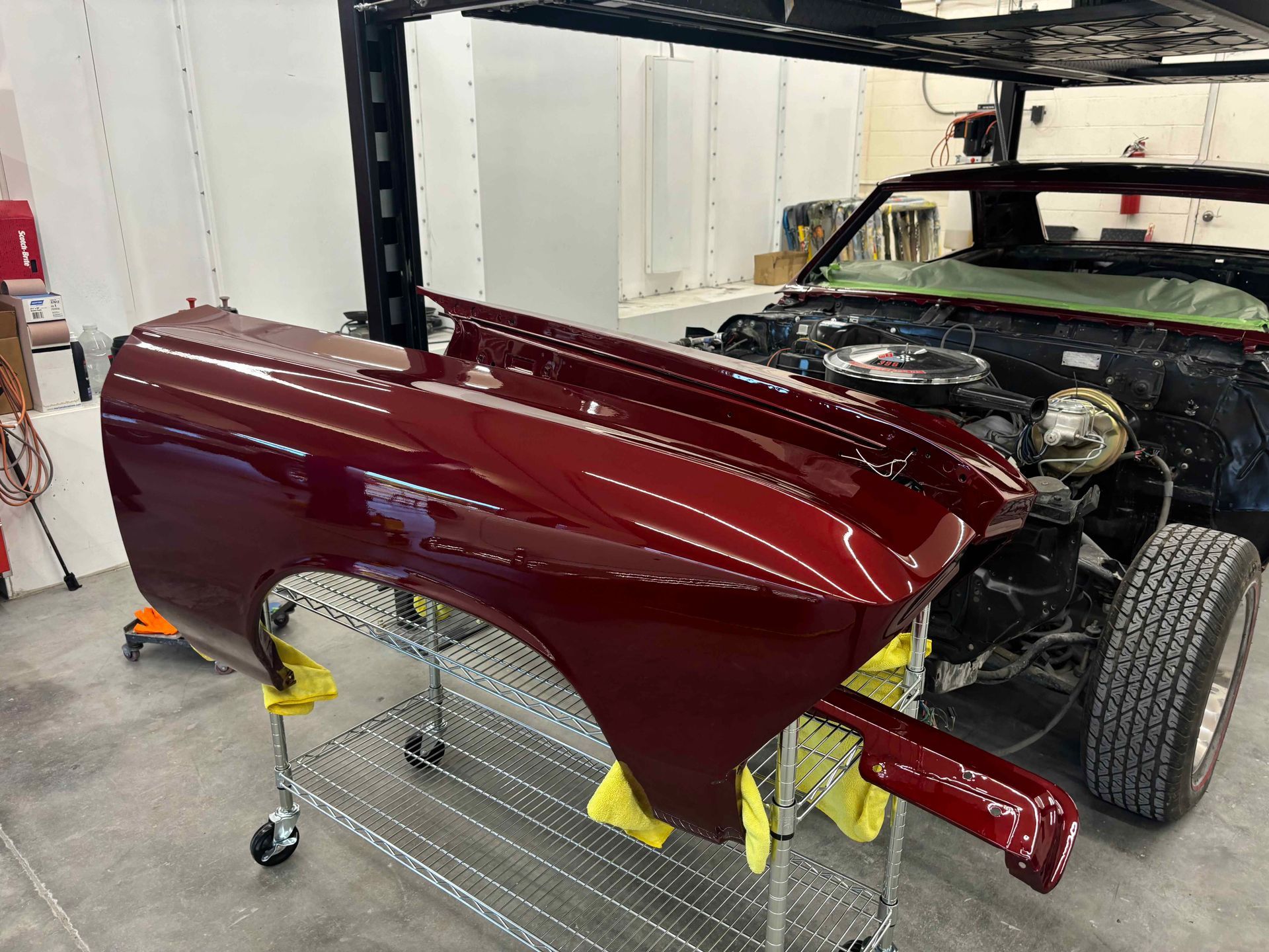Maroon car body shell on a stand in a workshop beside a black vehicle chassis