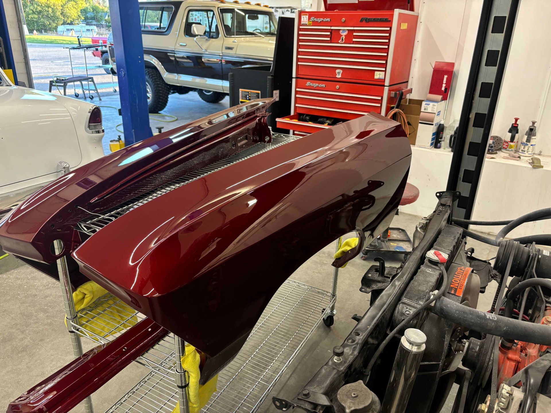 Maroon car body panels in an auto body shop with a red tool chest and workshop equipment.