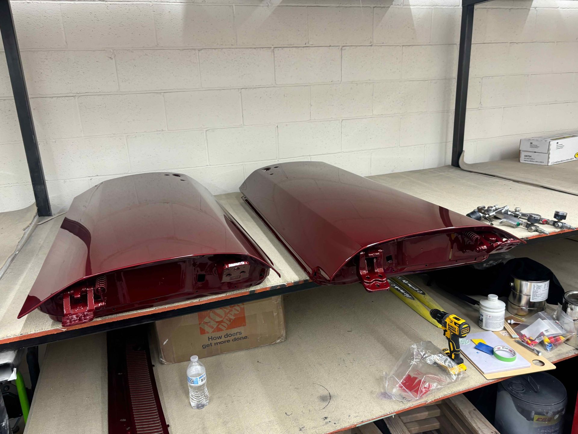 Two maroon car parts on a workbench in a garage, with tools and supplies nearby.