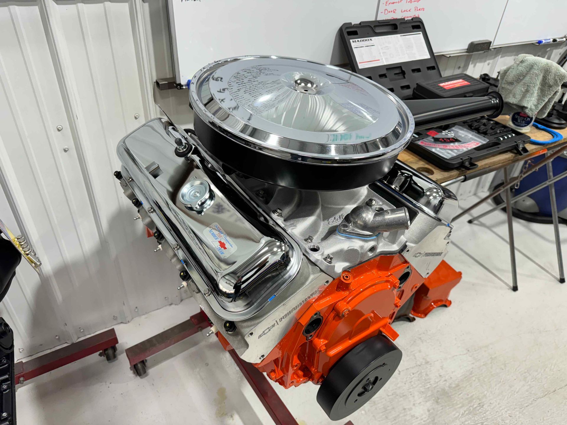 Shiny V8 engine on a stand in a workshop, with orange front cover and silver air cleaner.