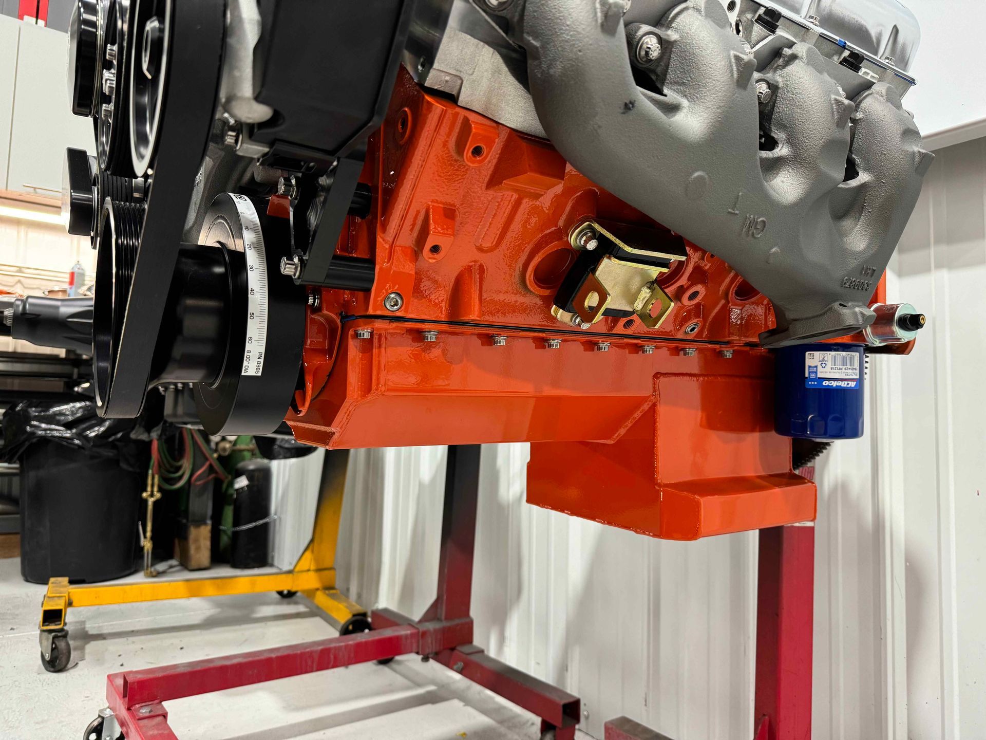 Orange engine block mounted on a stand in a workshop, with gray engine parts above it