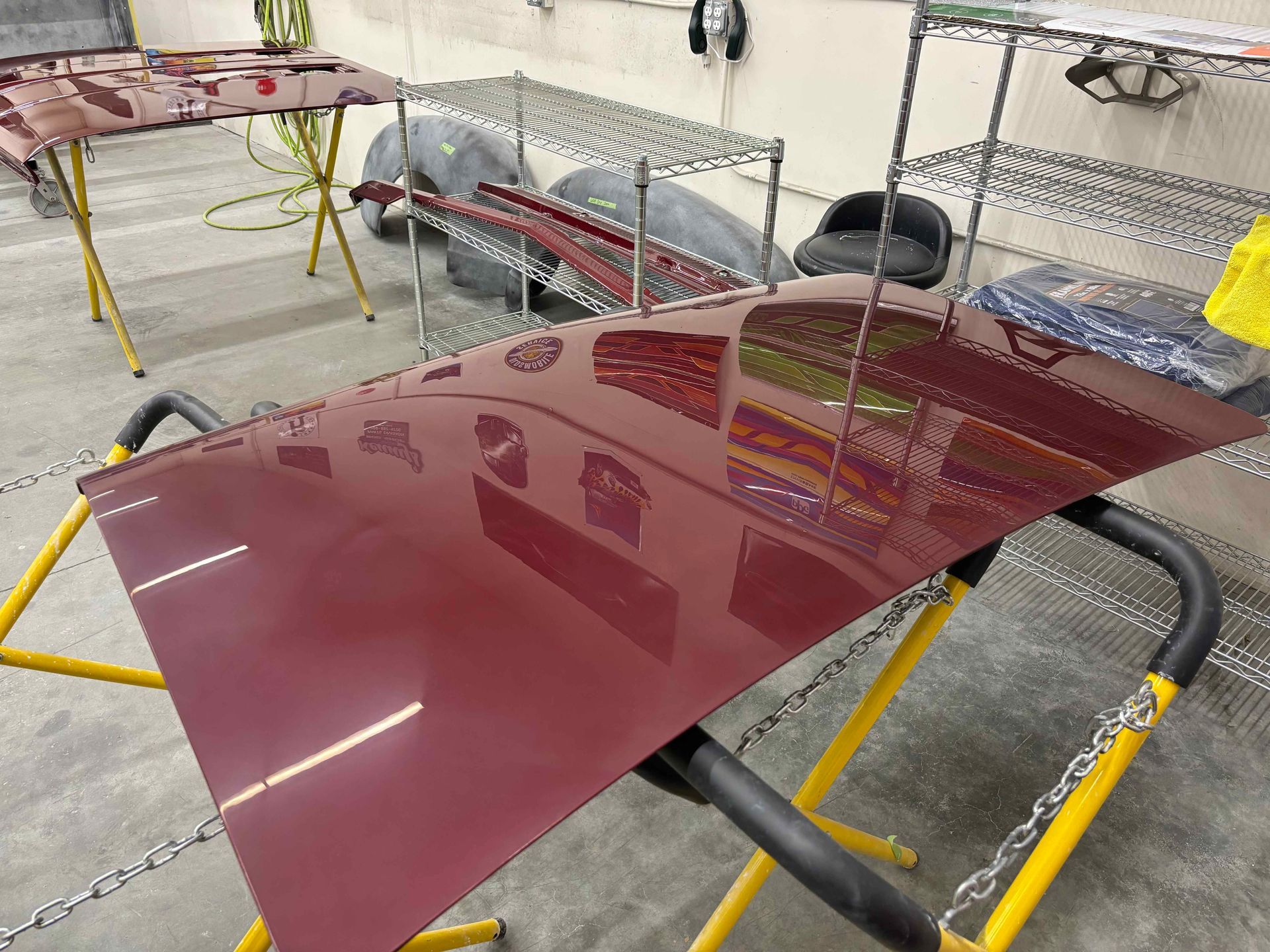 Glossy maroon car hood panel on a stand in an auto body shop, with other parts in the background