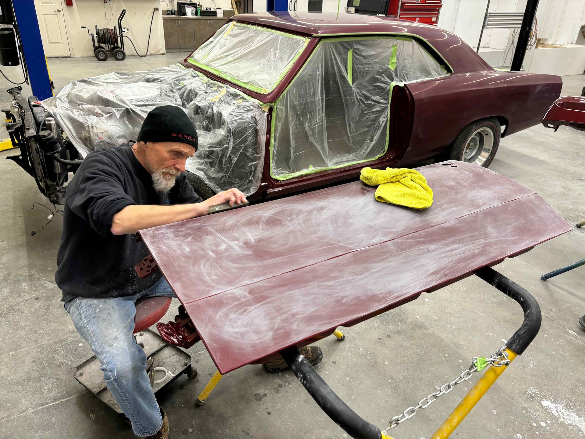 Worker sanding a maroon car body panel in a garage, with primer and plastic masking visible.