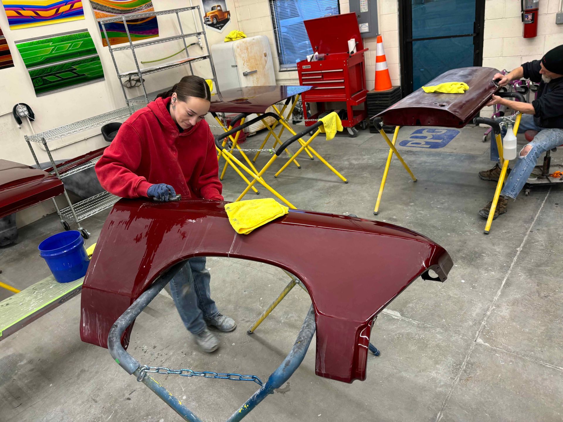 Person in a red sweater working on a maroon car panel in a workshop with other tools and tables nearby