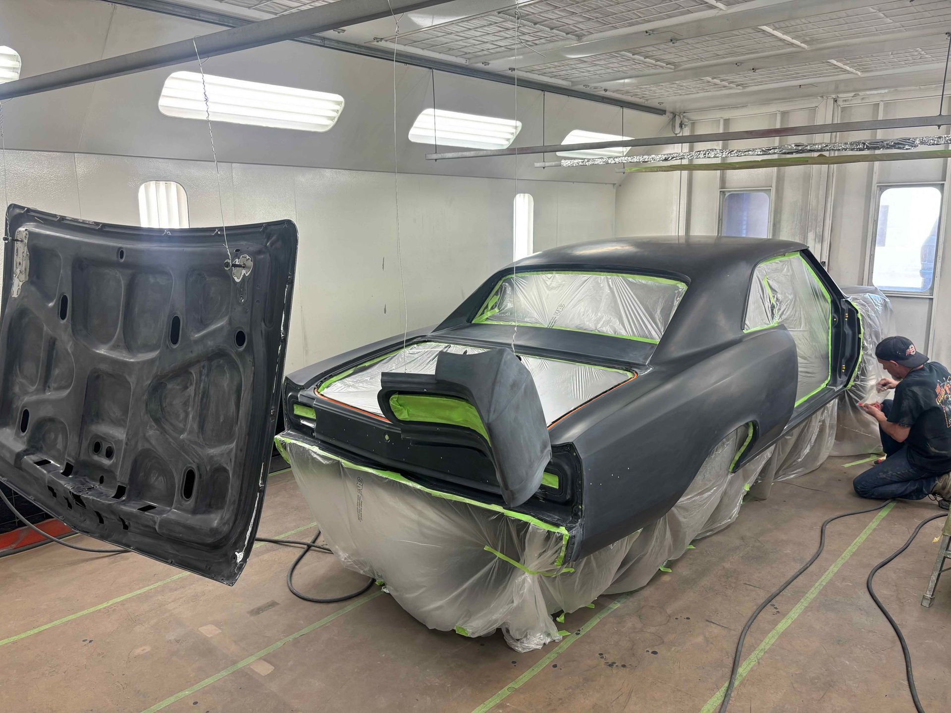 Car being painted in a body shop, masked in plastic with doors off and hood open; worker crouches nearby.