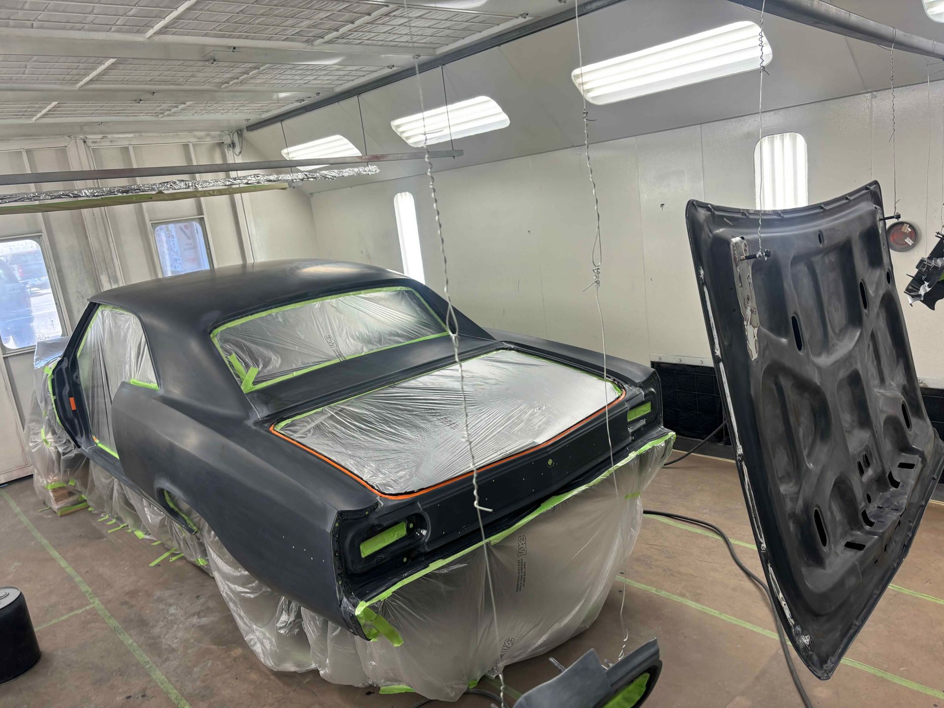 Classic car body primed for painting in a spray booth, with hood propped open nearby.