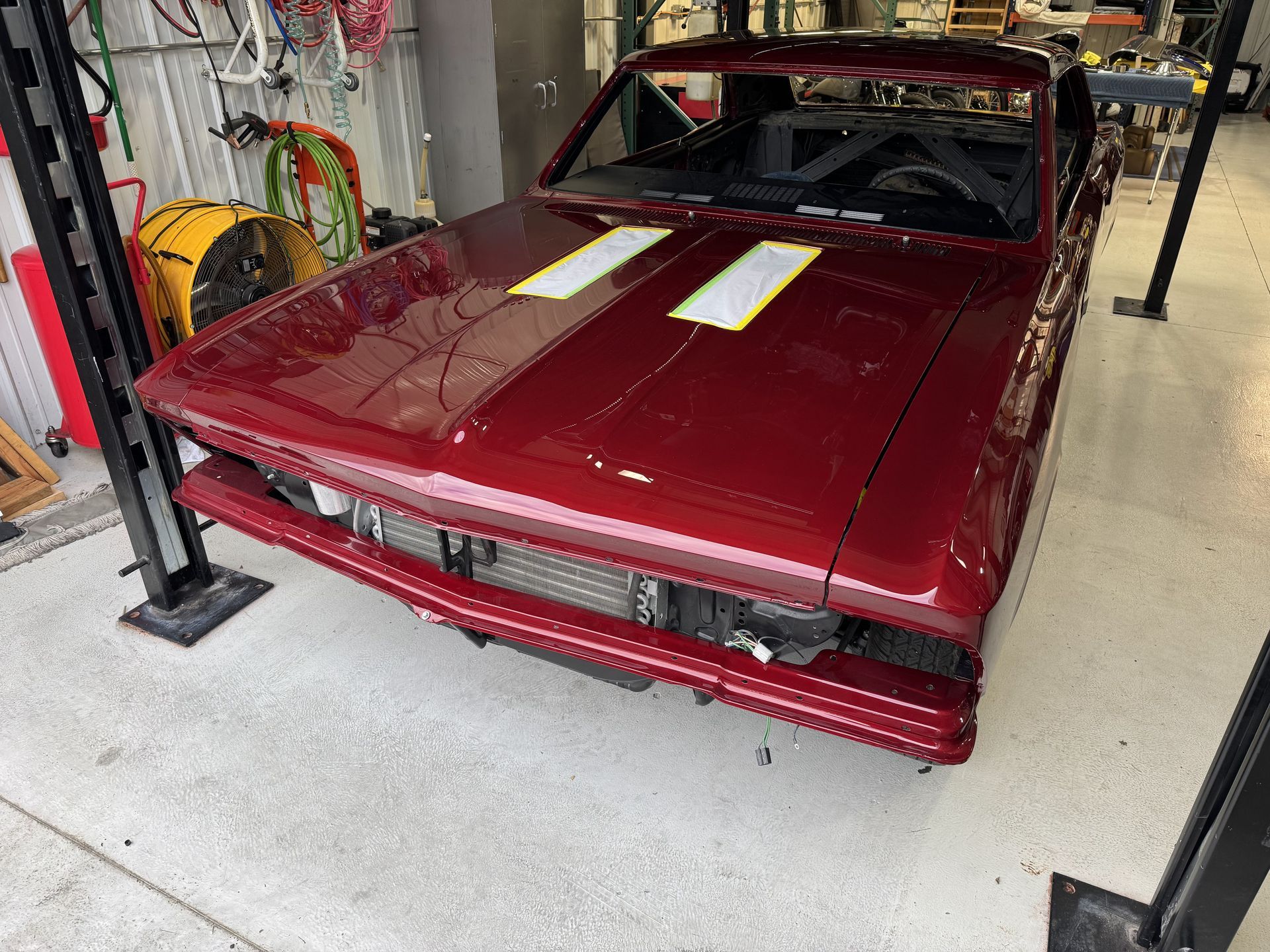 Maroon classic car in an auto shop, viewed from the front with hood stripes and bumper removed