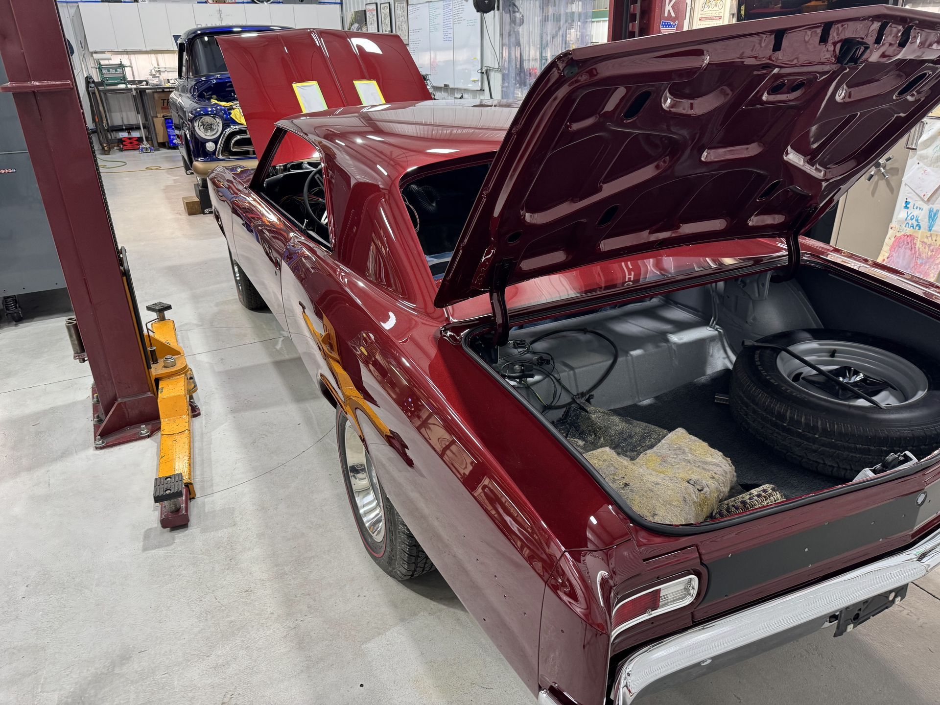 Maroon classic car in a workshop with hood and trunk open, showing the engine bay and rear storage.