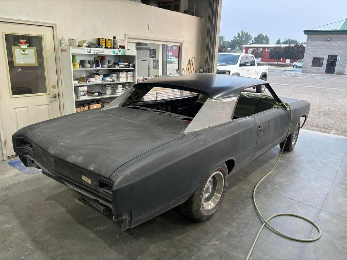 Primered classic car body in a garage, with shelves and a hose on the floor.