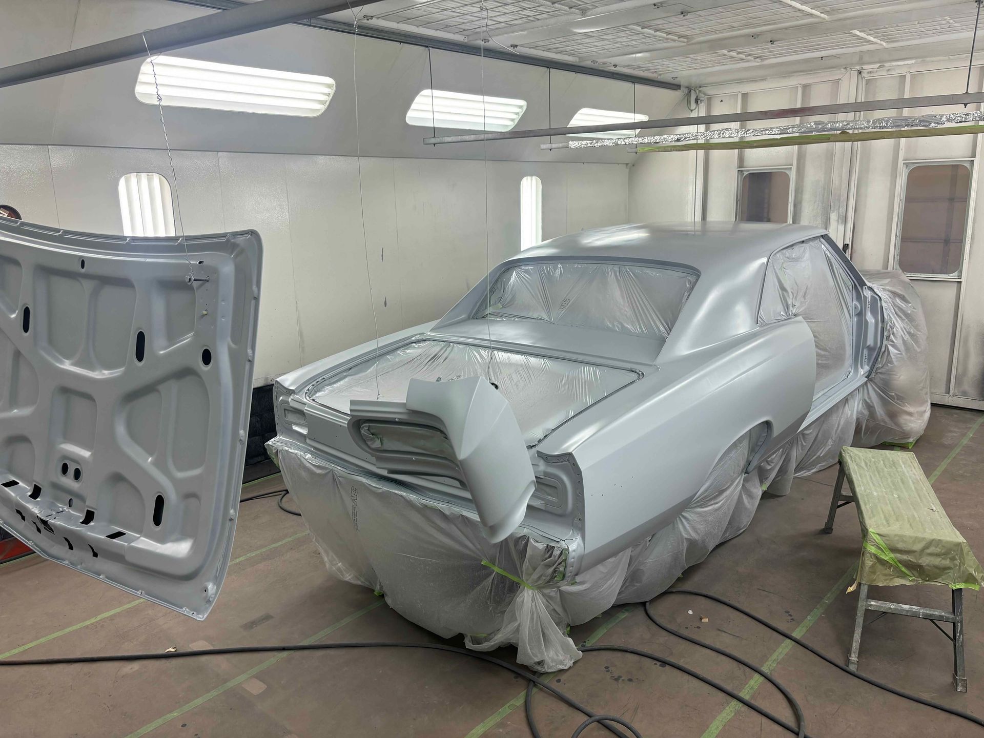 Car masked in primer inside a paint booth, with detached hood and covered windows.