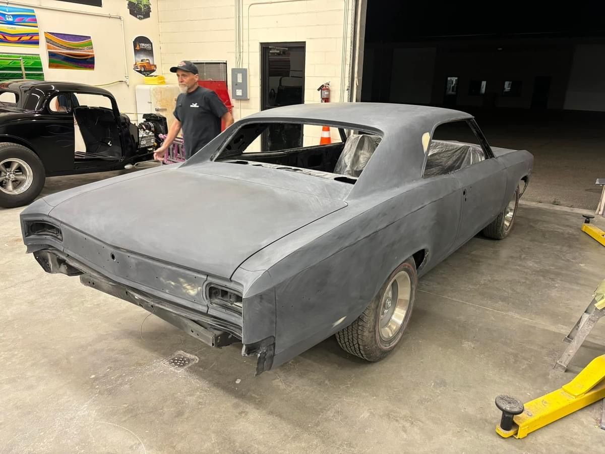 Gray classic car in a workshop, partially restored and covered in primer, with a person standing nearby.