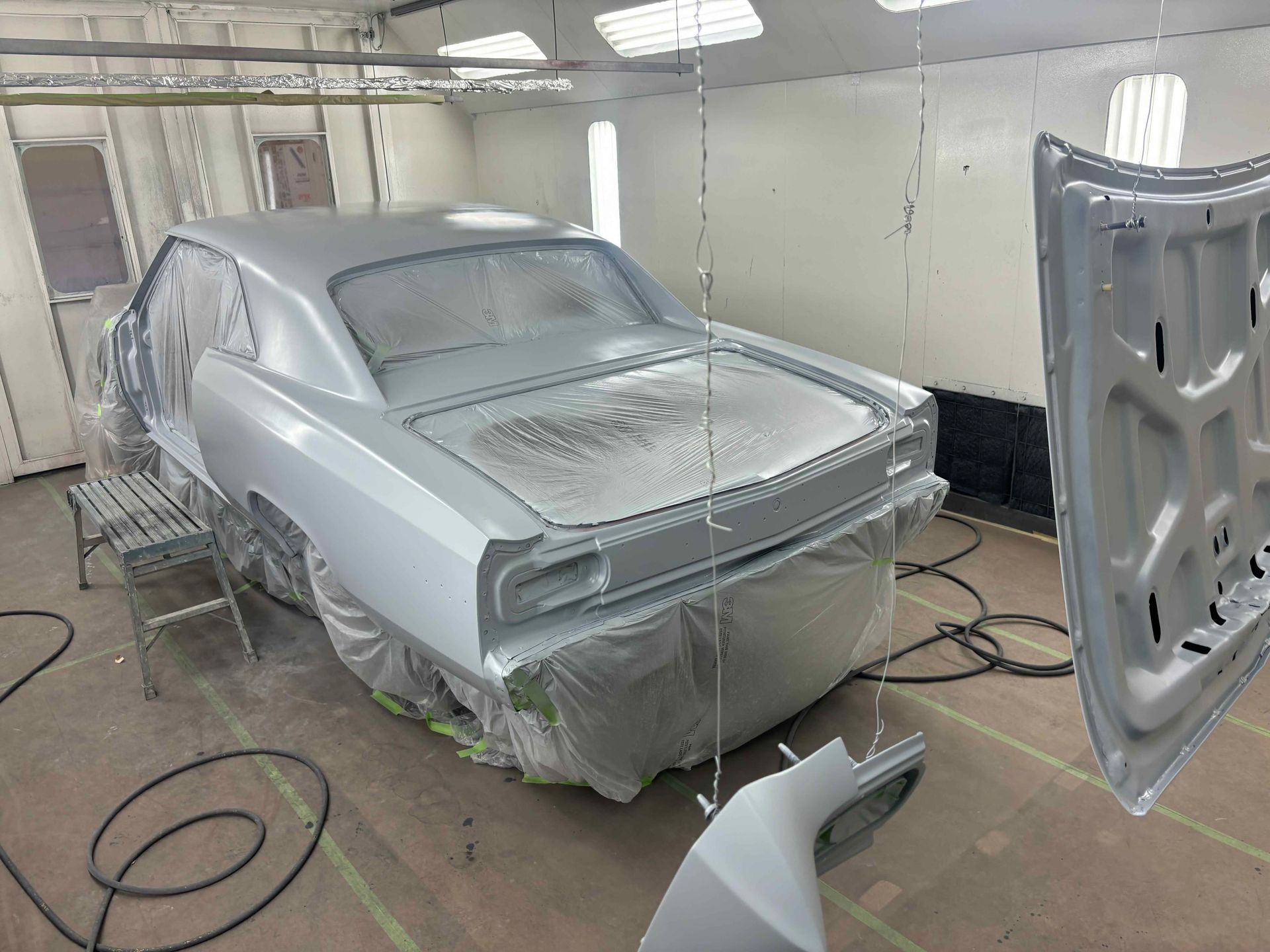 Silver car body in a paint booth, covered in plastic masking and primer, with detached panels nearby.
