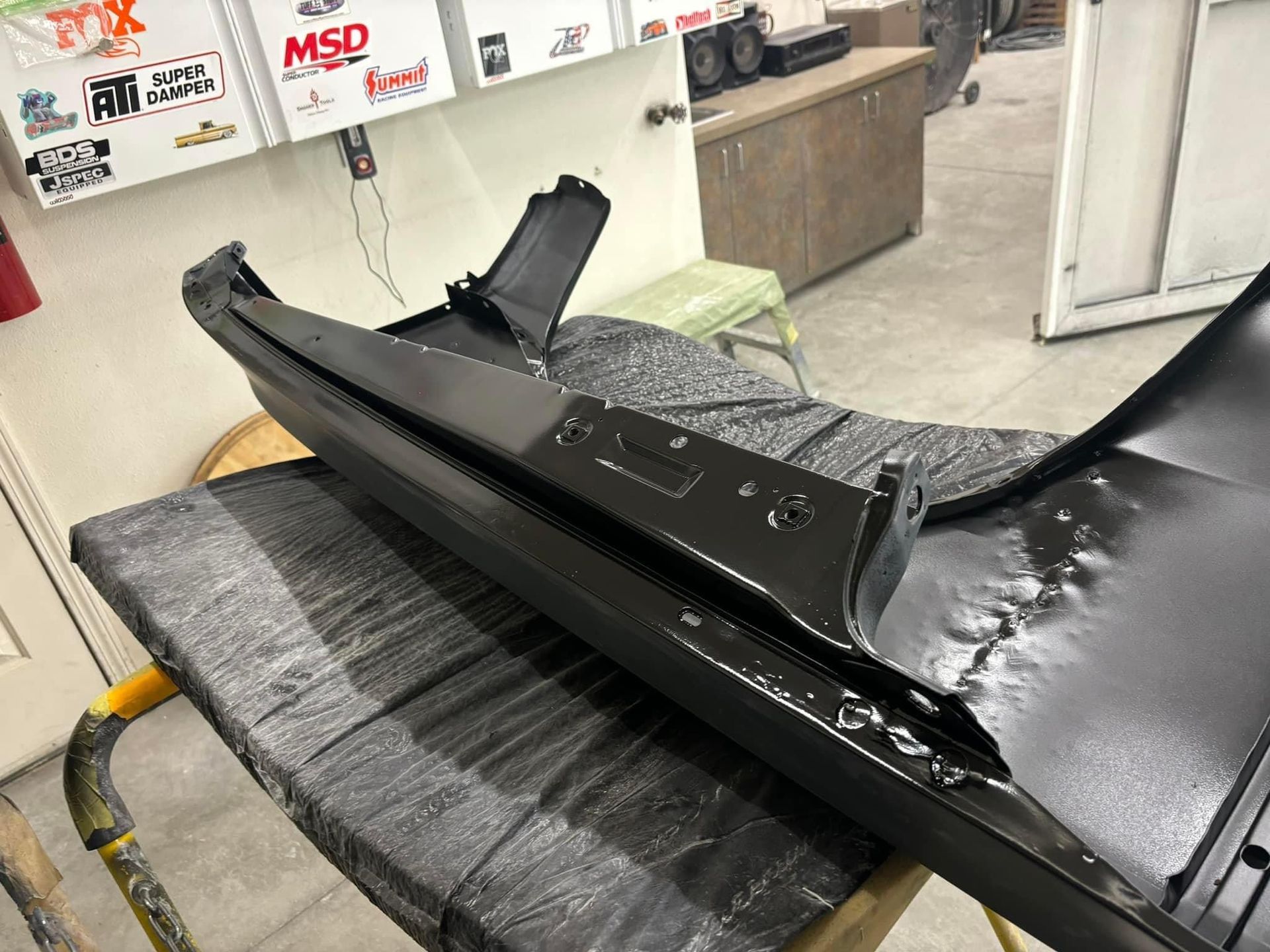 Black car bumper and body panel wrapped in protective film on a workbench in an auto shop