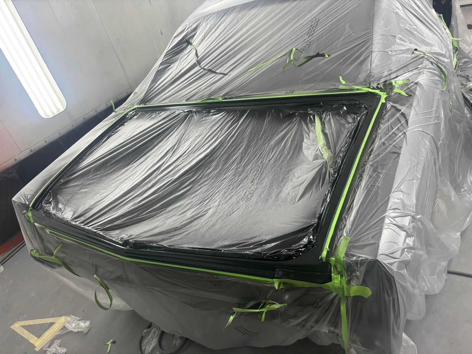 Car covered in gray plastic and green tape inside a paint booth