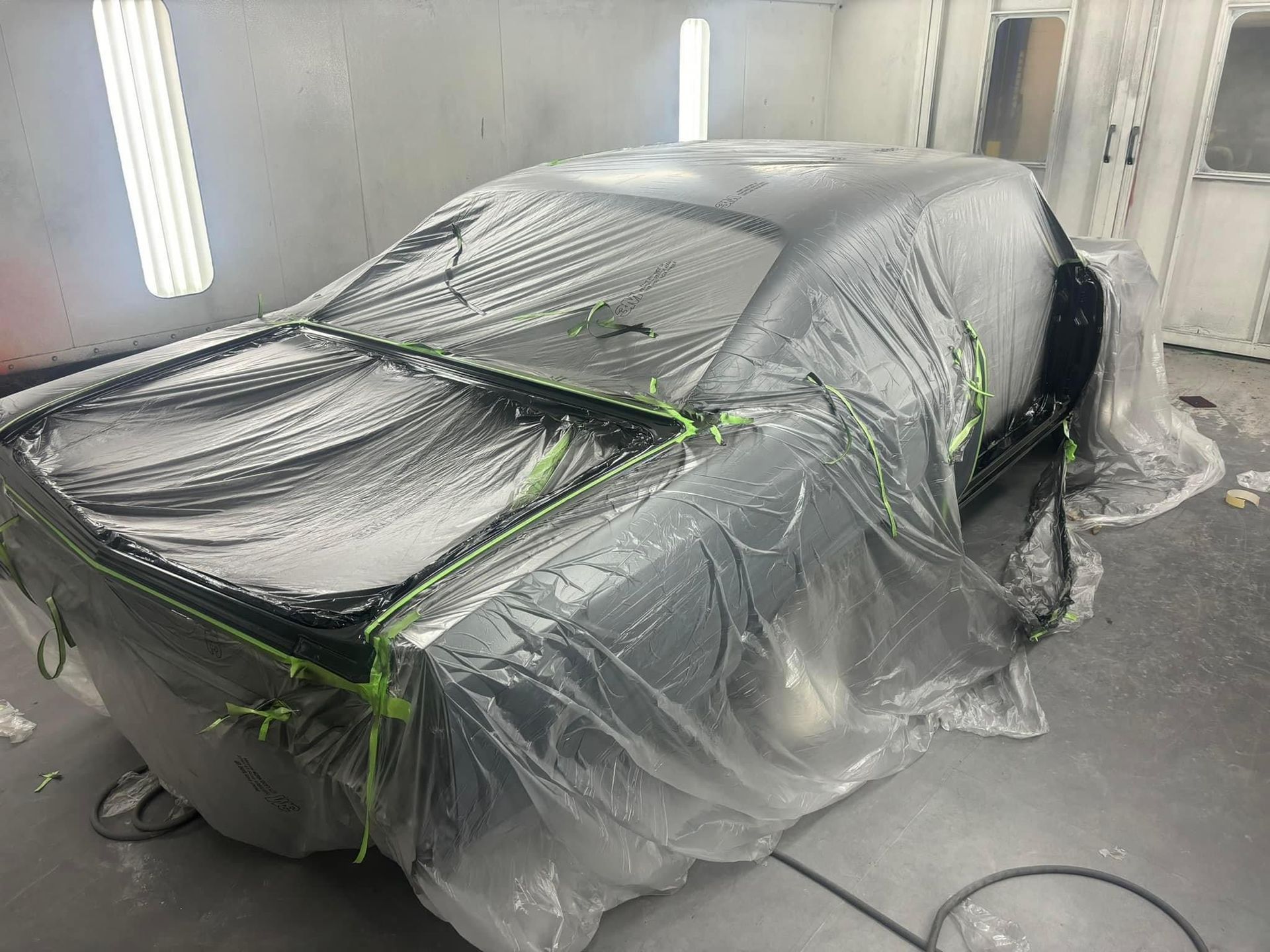 Black car body wrapped in plastic in a paint booth, with green masking tape around edges