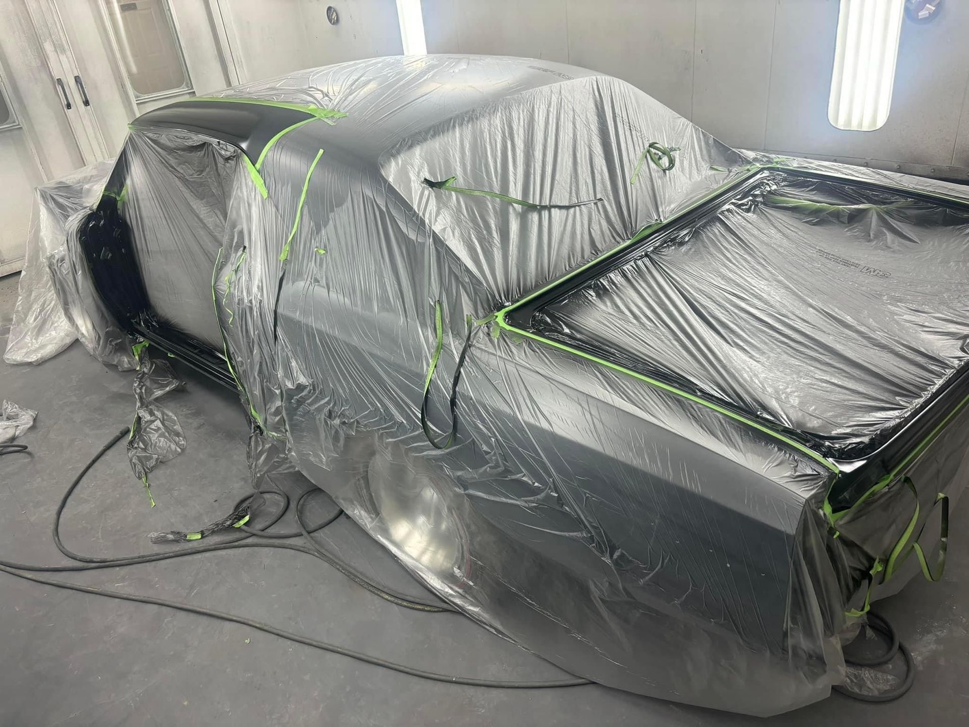 Car wrapped in plastic sheeting in a body shop, with windows masked for painting.