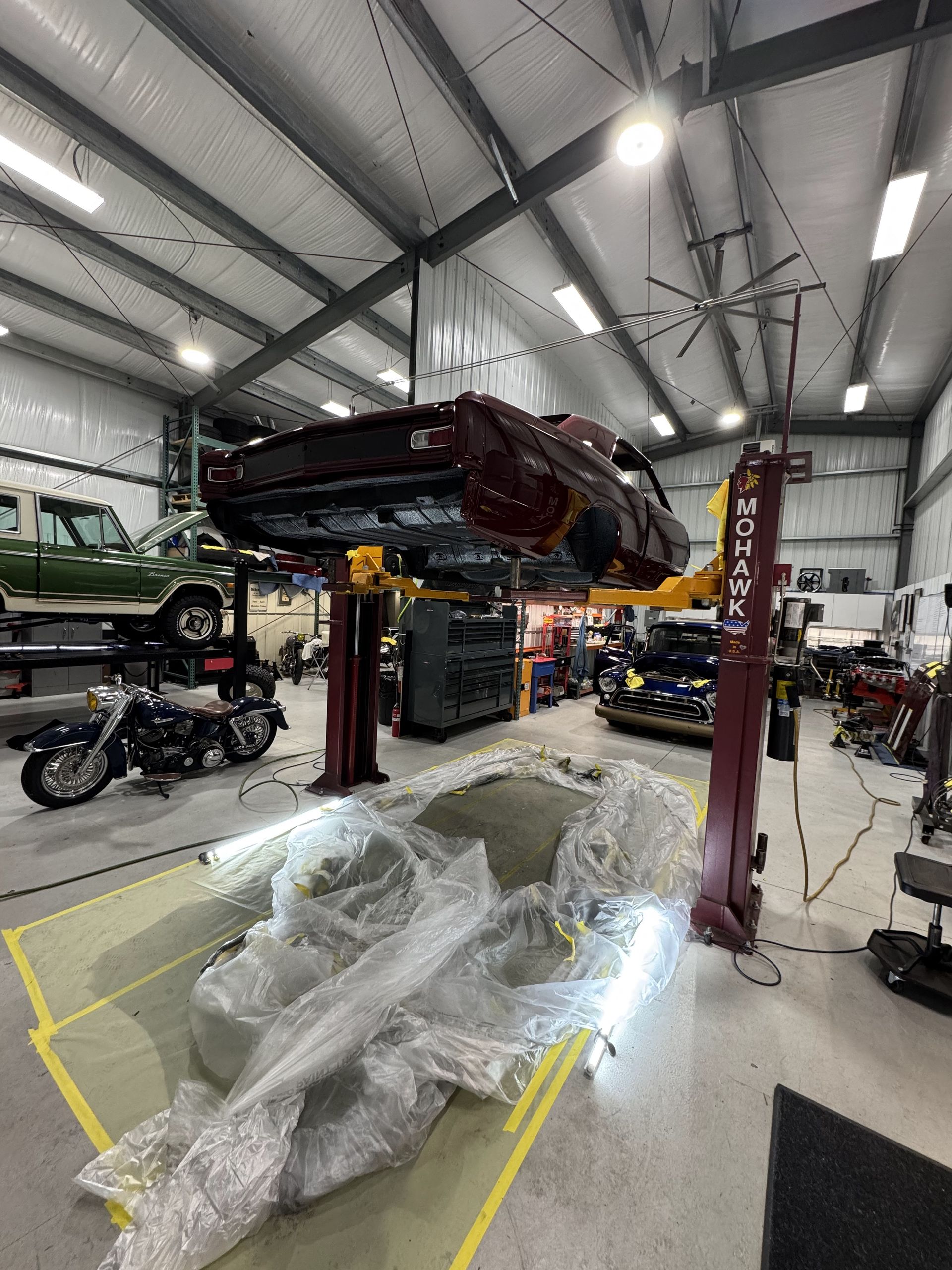 Car lifted on a garage hoist, with a covered engine or body panel on the floor below.