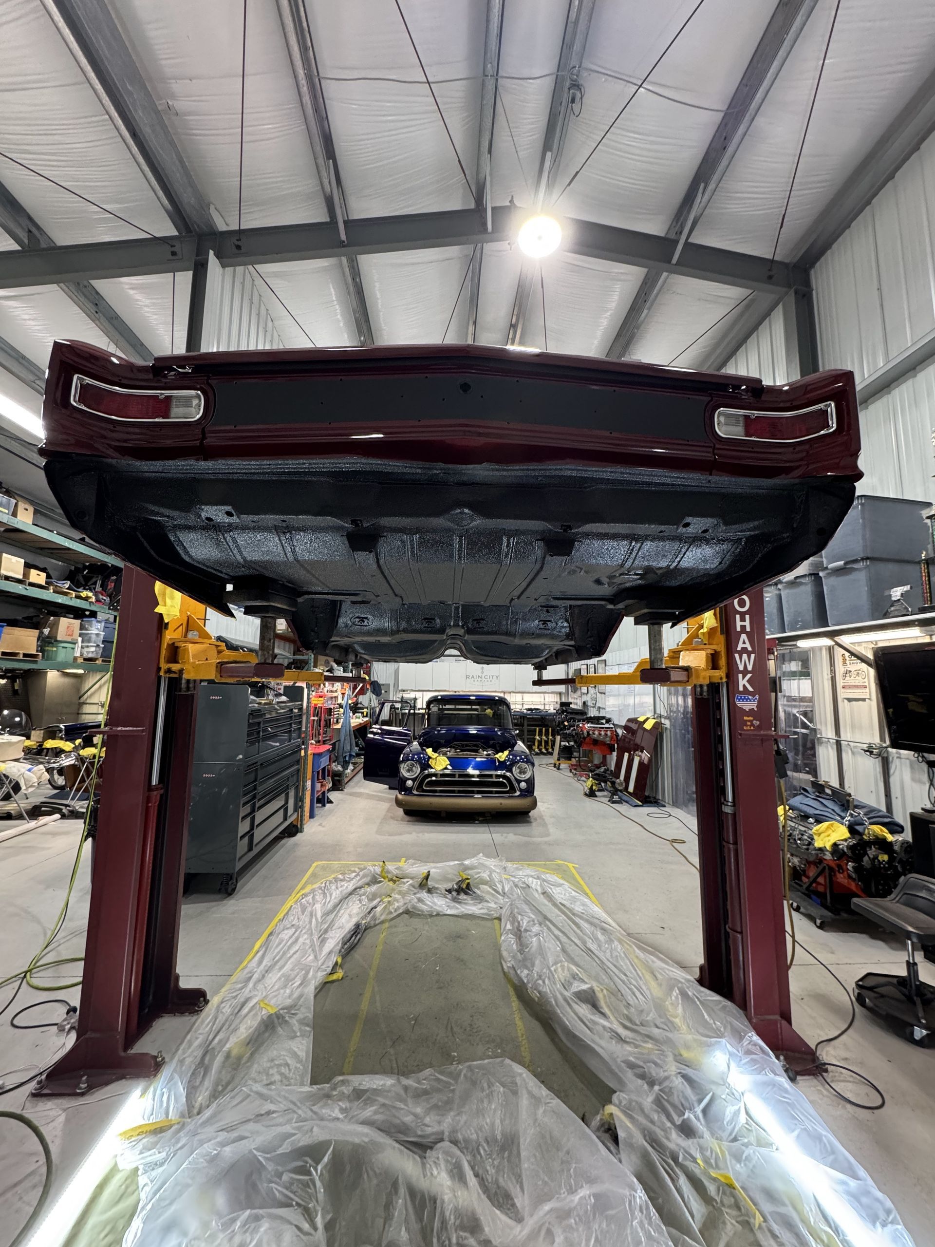 Car on a lift in a mechanic’s shop, with its underside exposed and a covered car below.