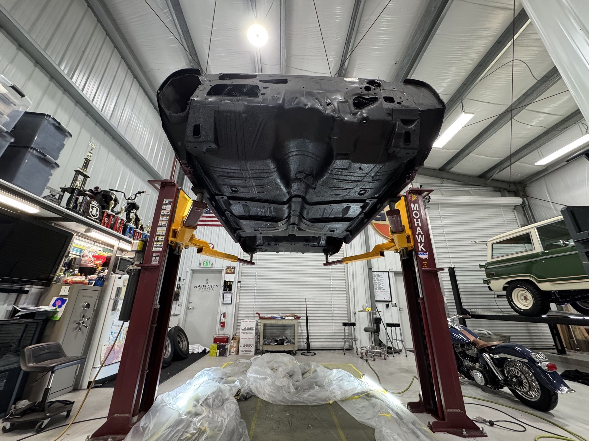 Car lifted on a shop hoist in an auto repair garage, underside visible with tools and vehicles nearby