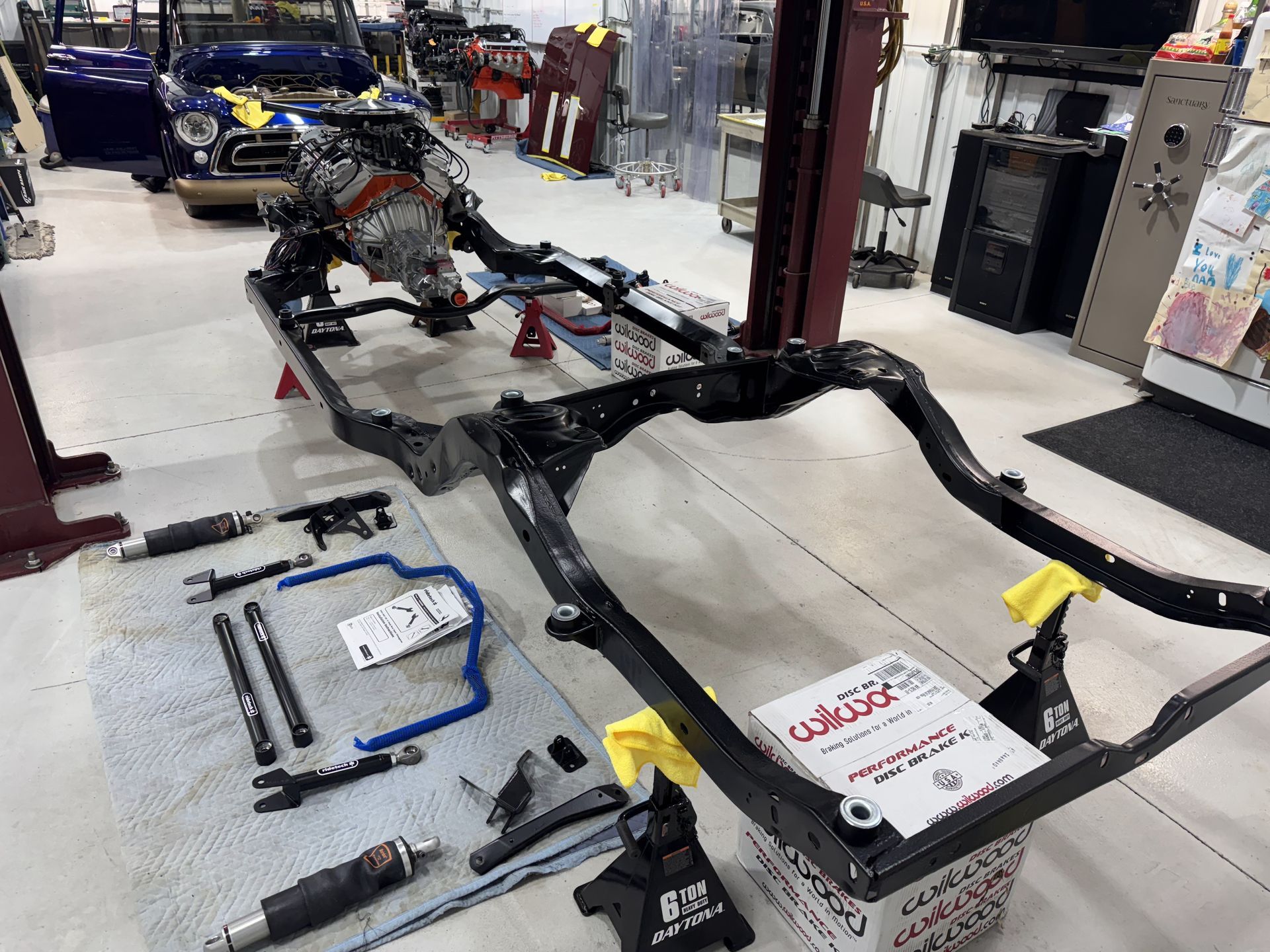 Vehicle chassis frame on workshop floor surrounded by tools and parts, with cars and equipment in the background