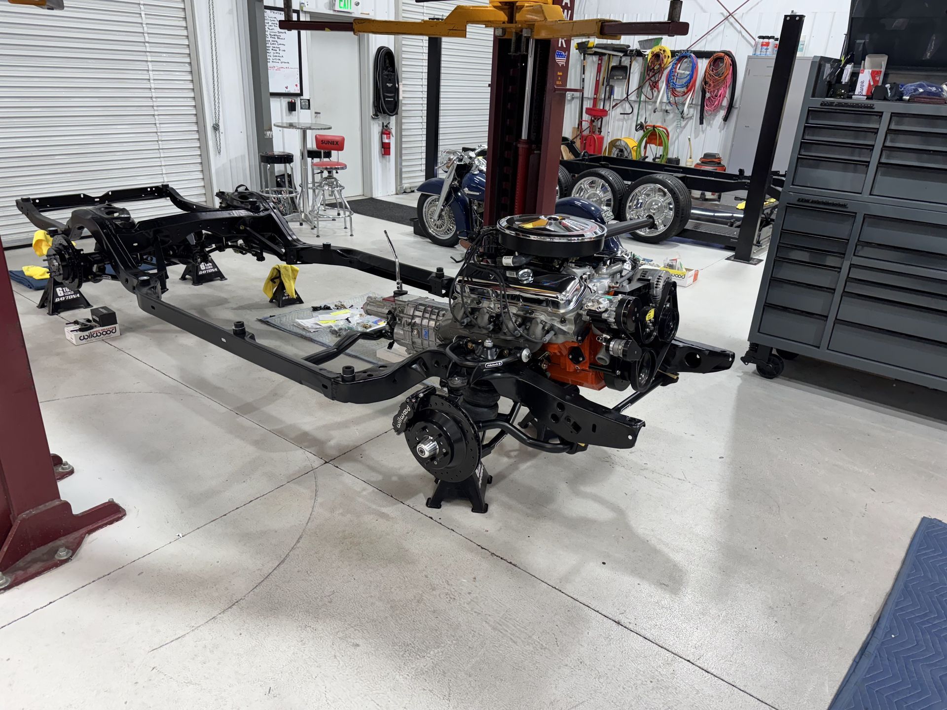Car chassis frame and engine assembly in a workshop, with tools and storage racks nearby.