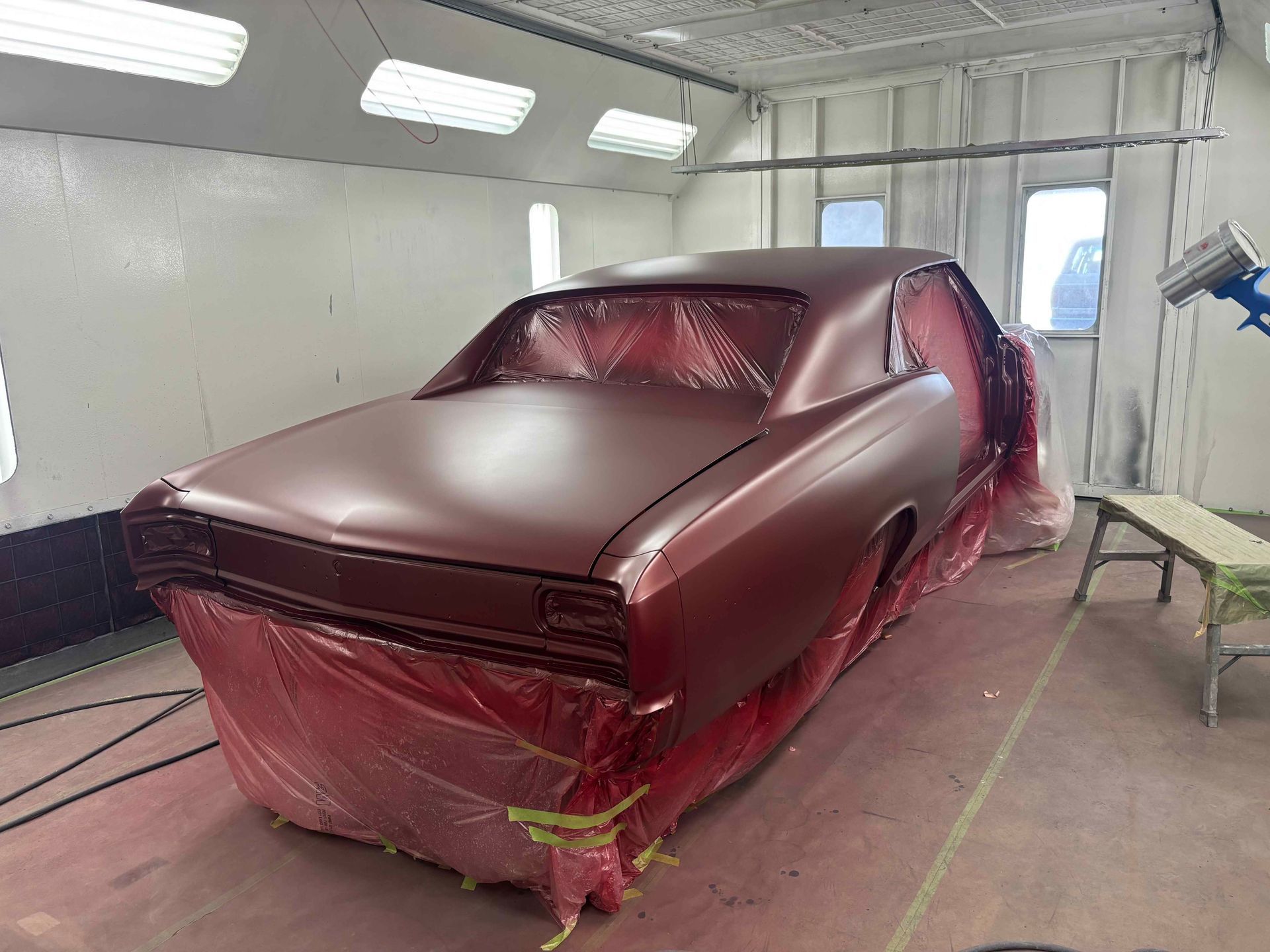 Maroon car body in a paint booth, masked with plastic sheeting and ready for painting