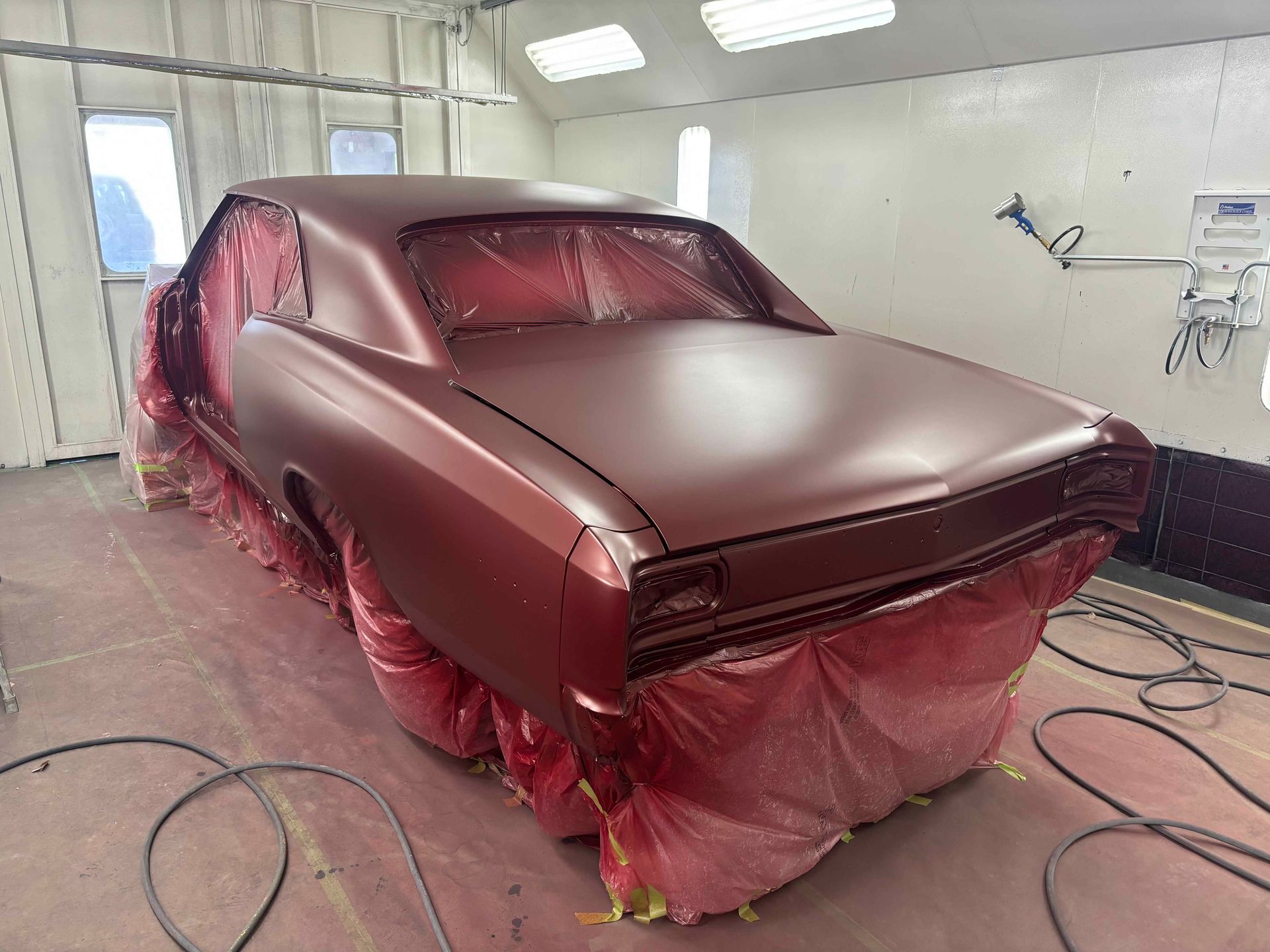 Car body being spray-painted in a workshop, covered in maroon primer and masked with plastic.