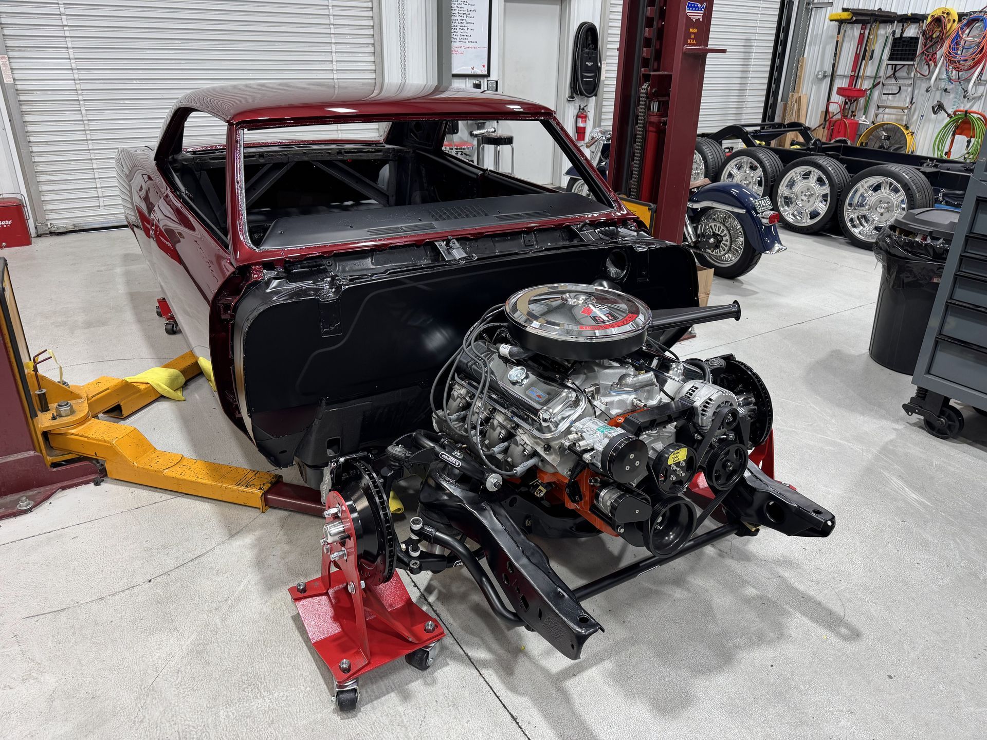 Classic car chassis and engine undergoing restoration in an auto shop, with body shell raised on a lift.