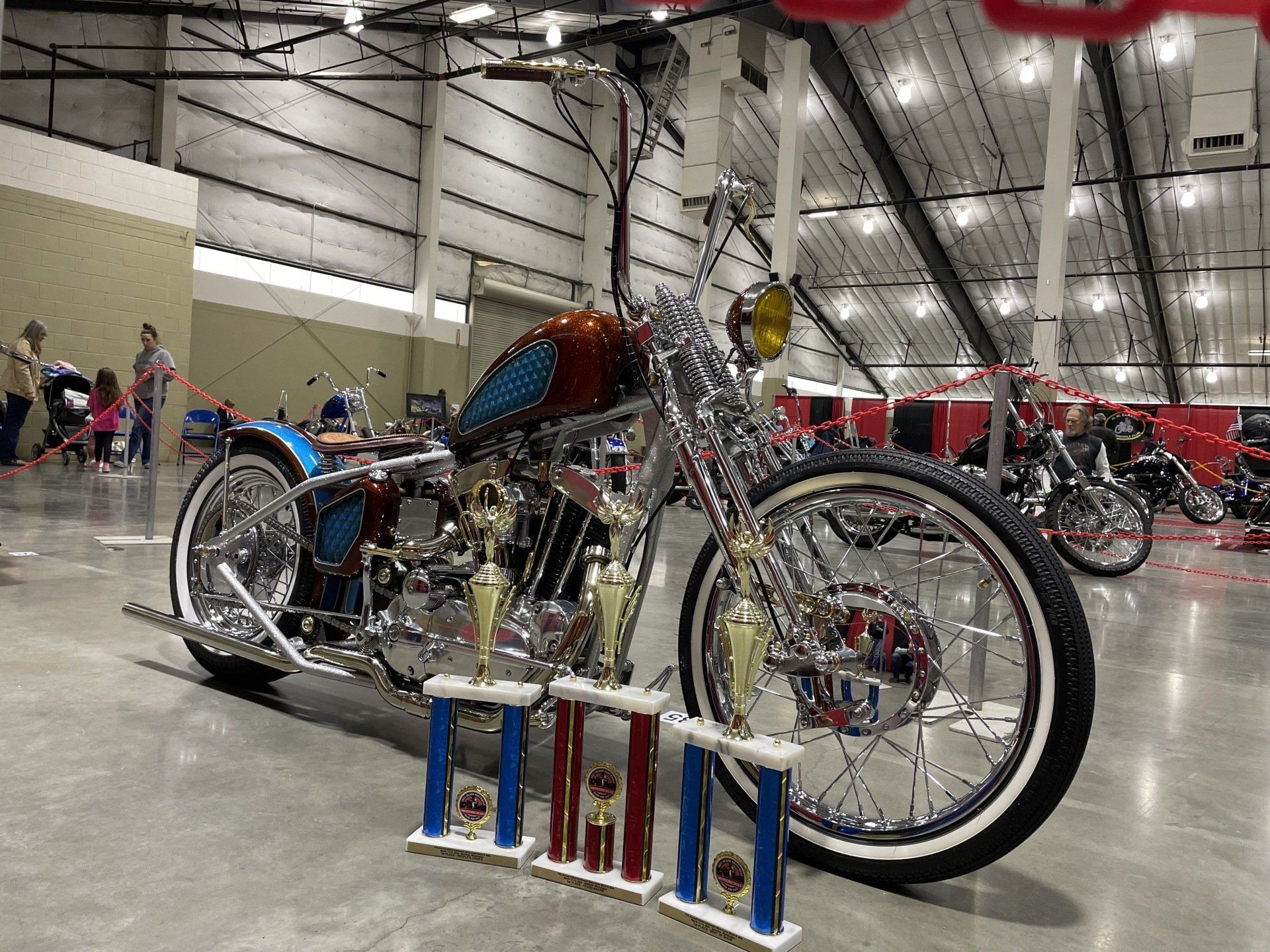 Custom chrome motorcycle on display indoors at a show, with trophies in front of it.