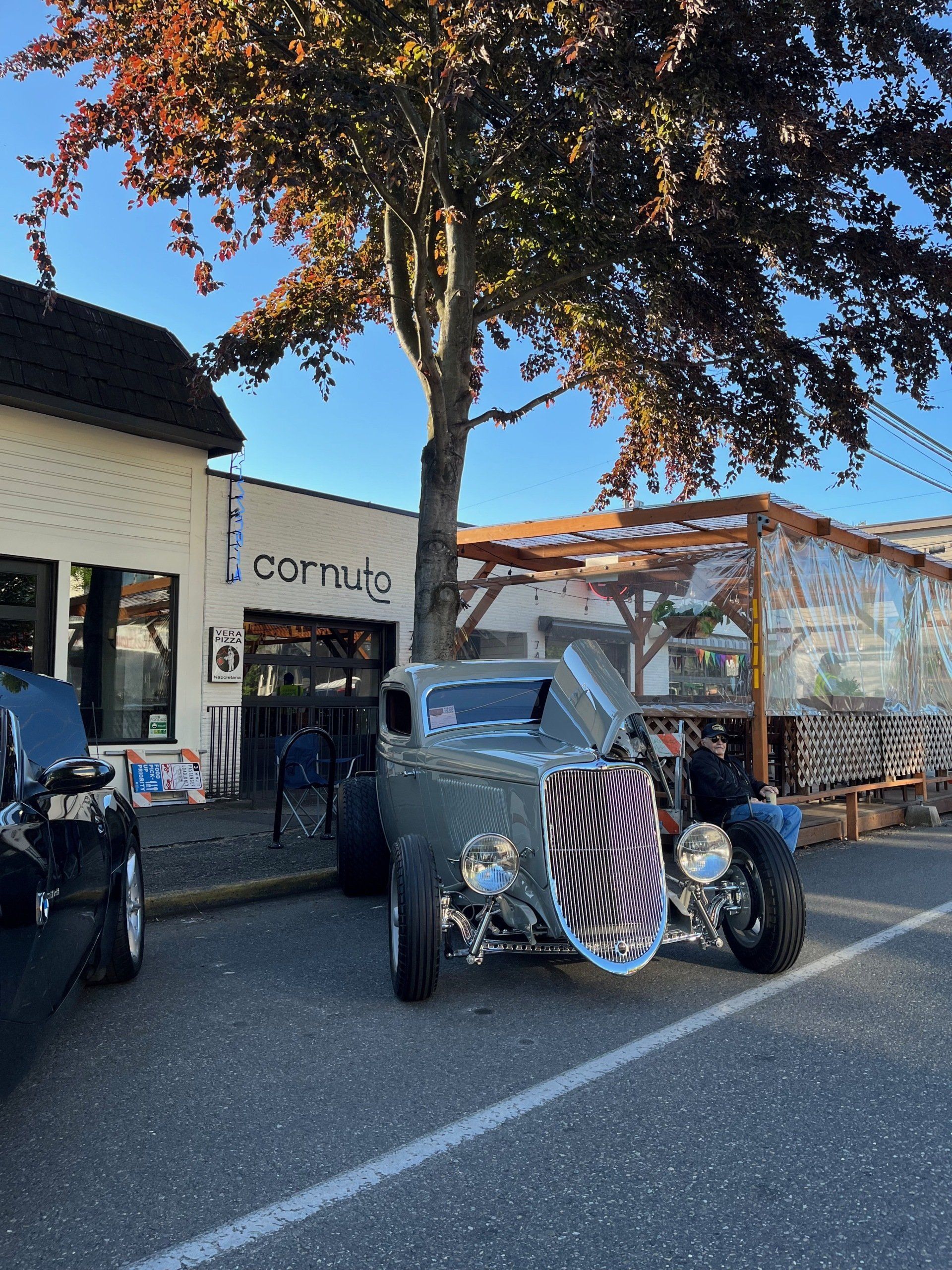 Vintage car parked outside a shop under a tree, with a blue truck beside it.