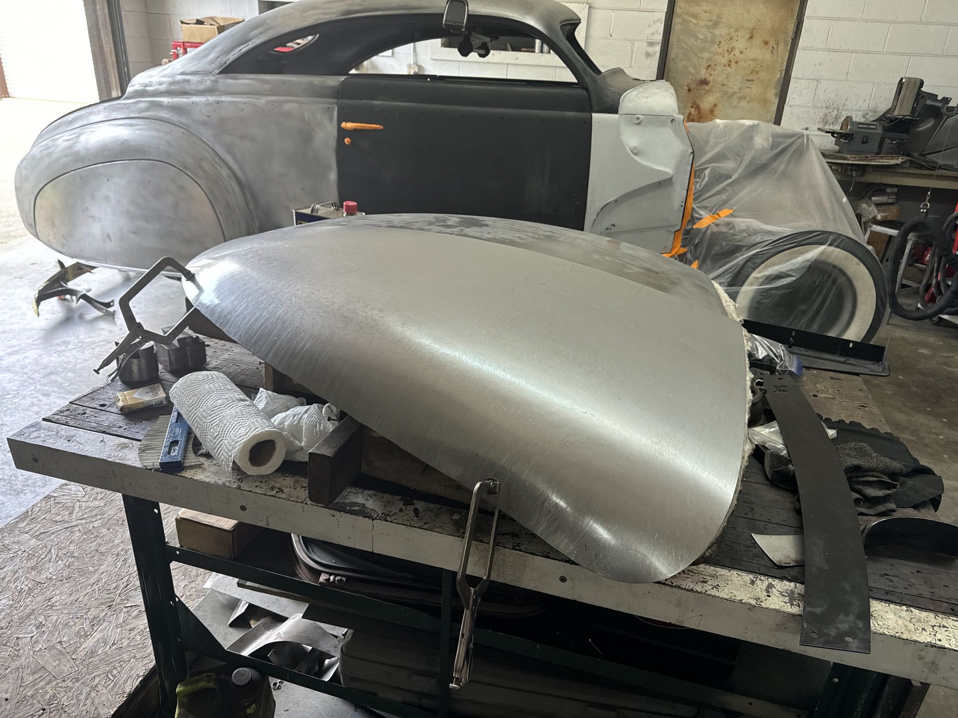 Silver car body parts and fenders stacked in a workshop, with a partially assembled vintage car in the background
