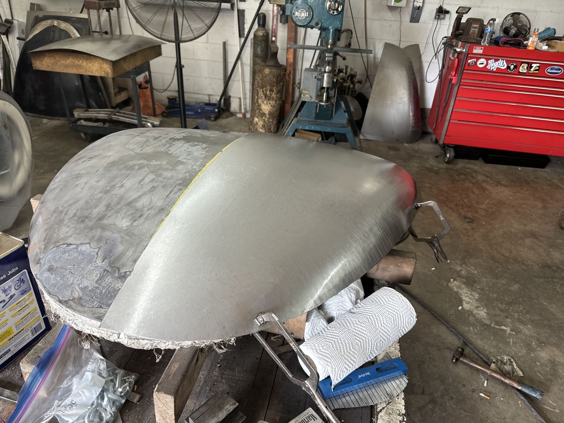 Car hood in a workshop, split between bare metal and gray primer, with tools and a red tool chest nearby